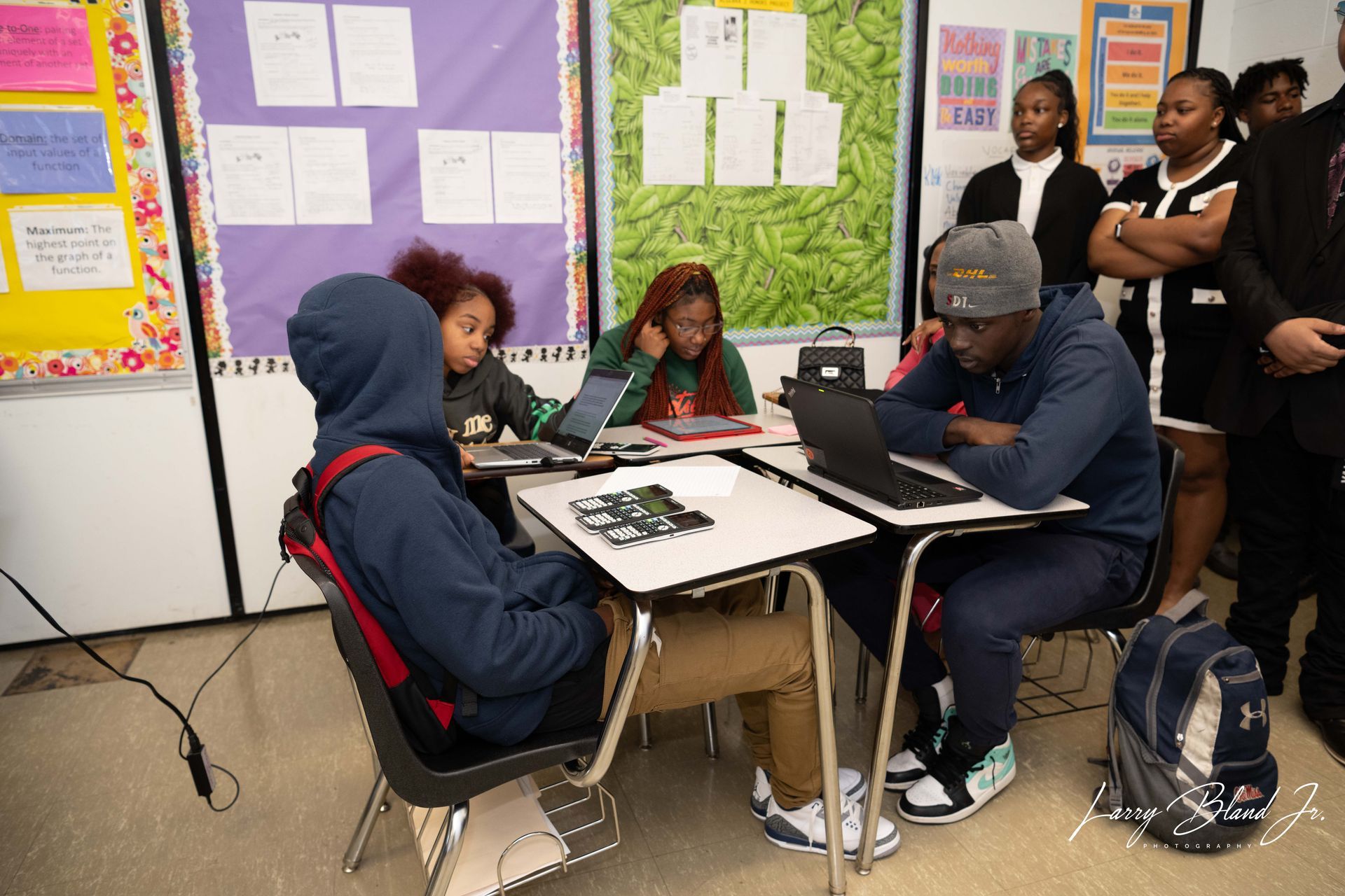 Students work at desks in a classroom with adult observers. One student has a laptop, others have tablets.