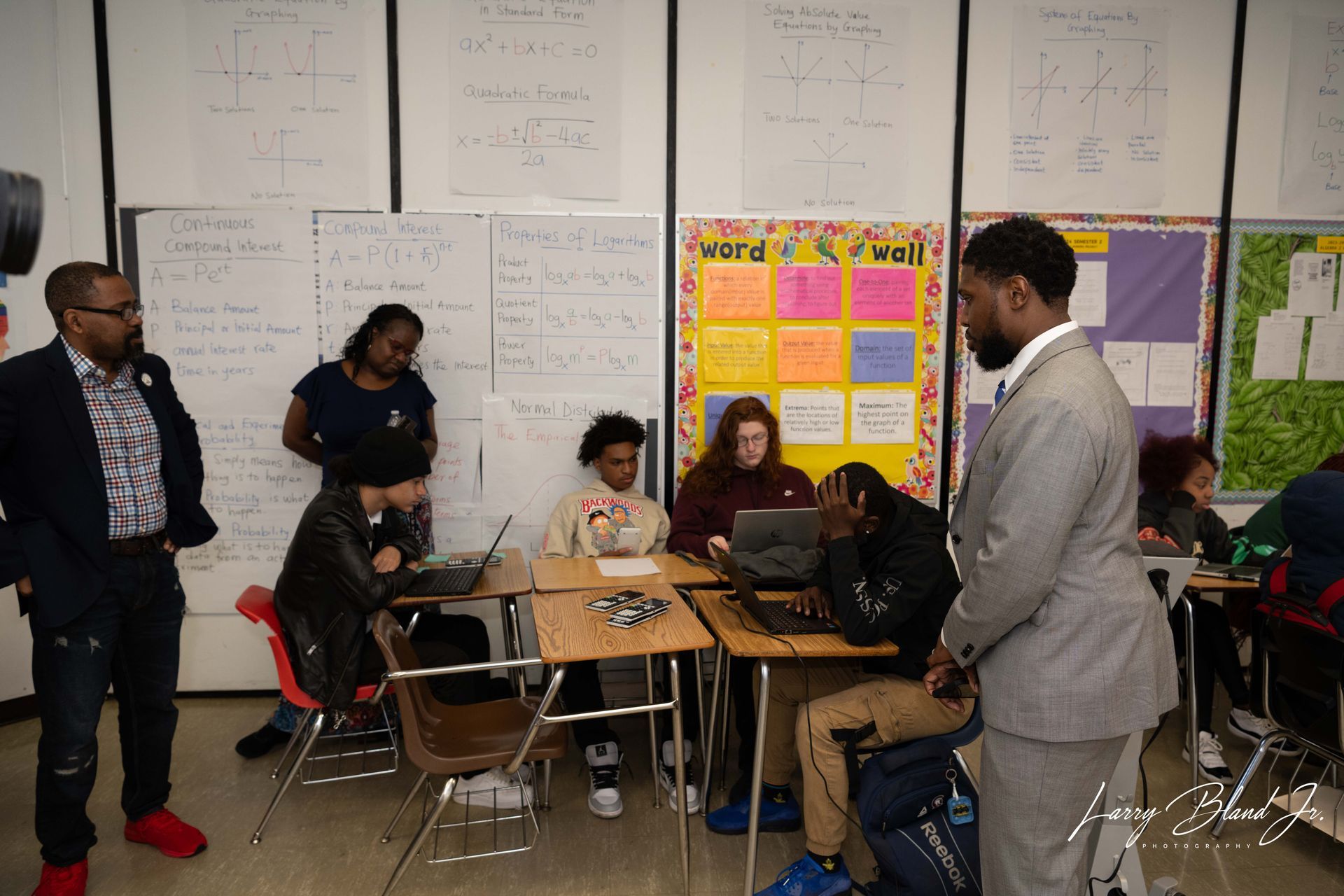 Classroom: Students at computers, teachers observing. Whiteboard with writing, colorful bulletin board.