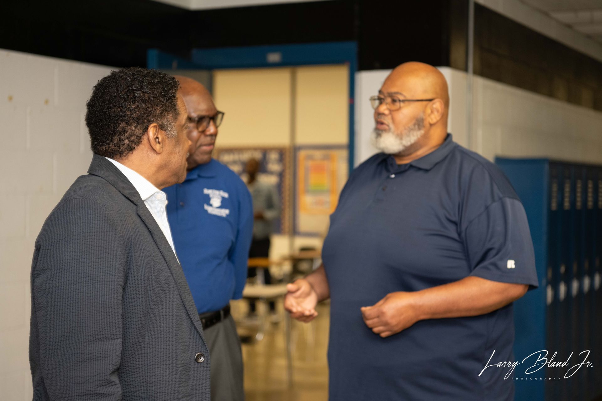 Three men conversing in a hallway with lockers. One in a blazer, one in a blue polo, one with a gray beard.