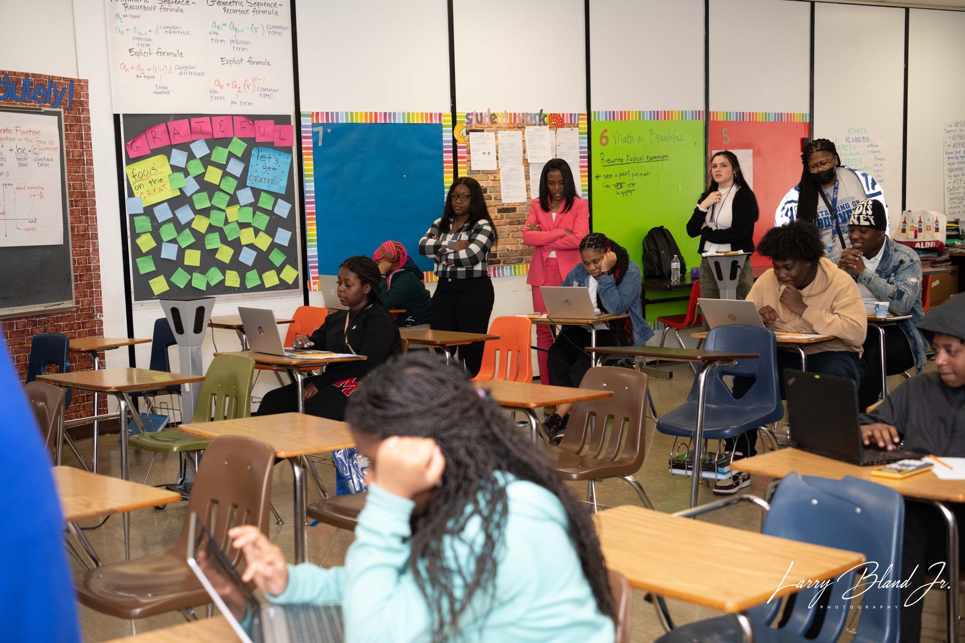 Classroom with students at desks using laptops, some standing in the back. Whiteboards, colorful posters on walls.