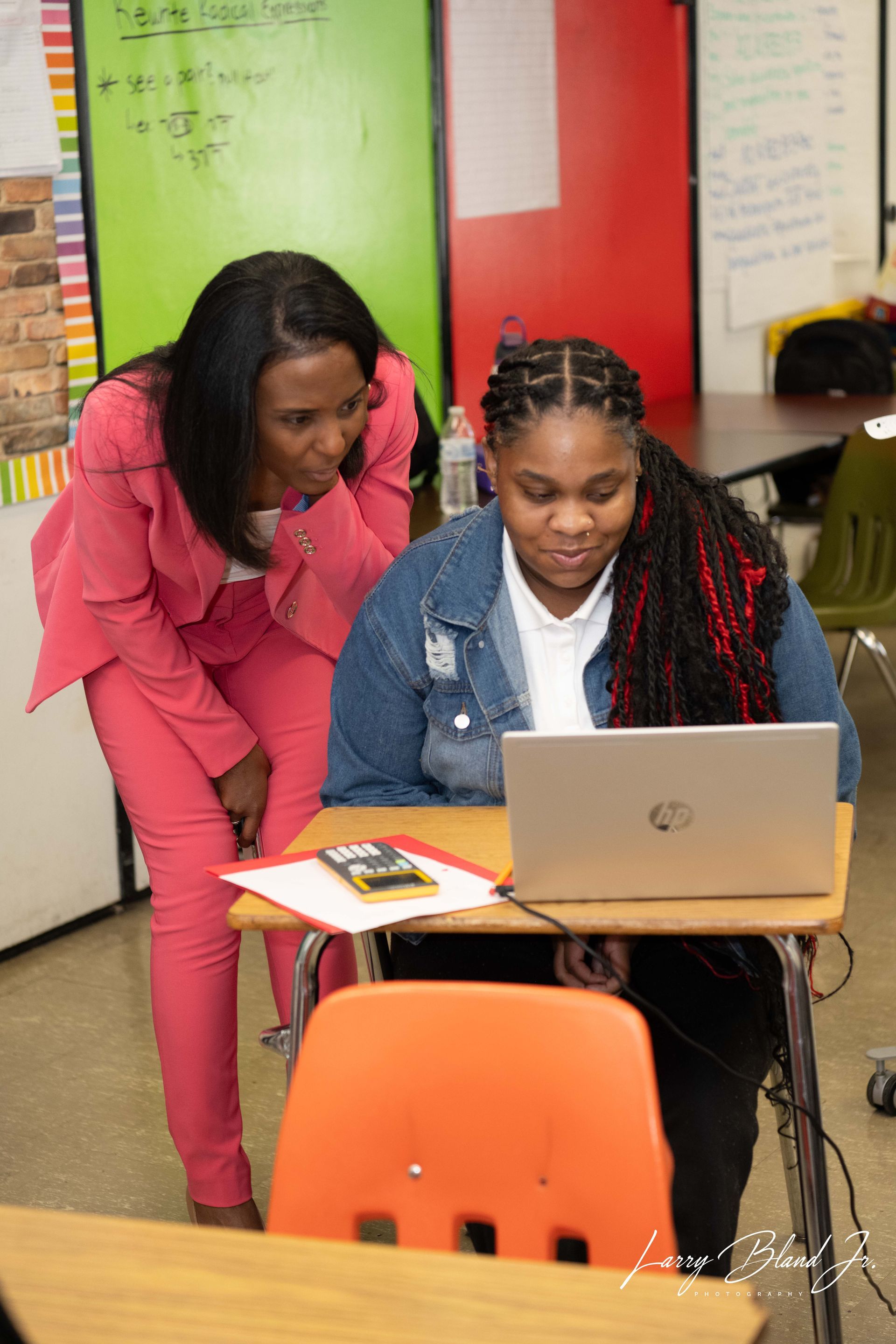 Woman in pink suit leans over student using a laptop at a desk in a classroom.