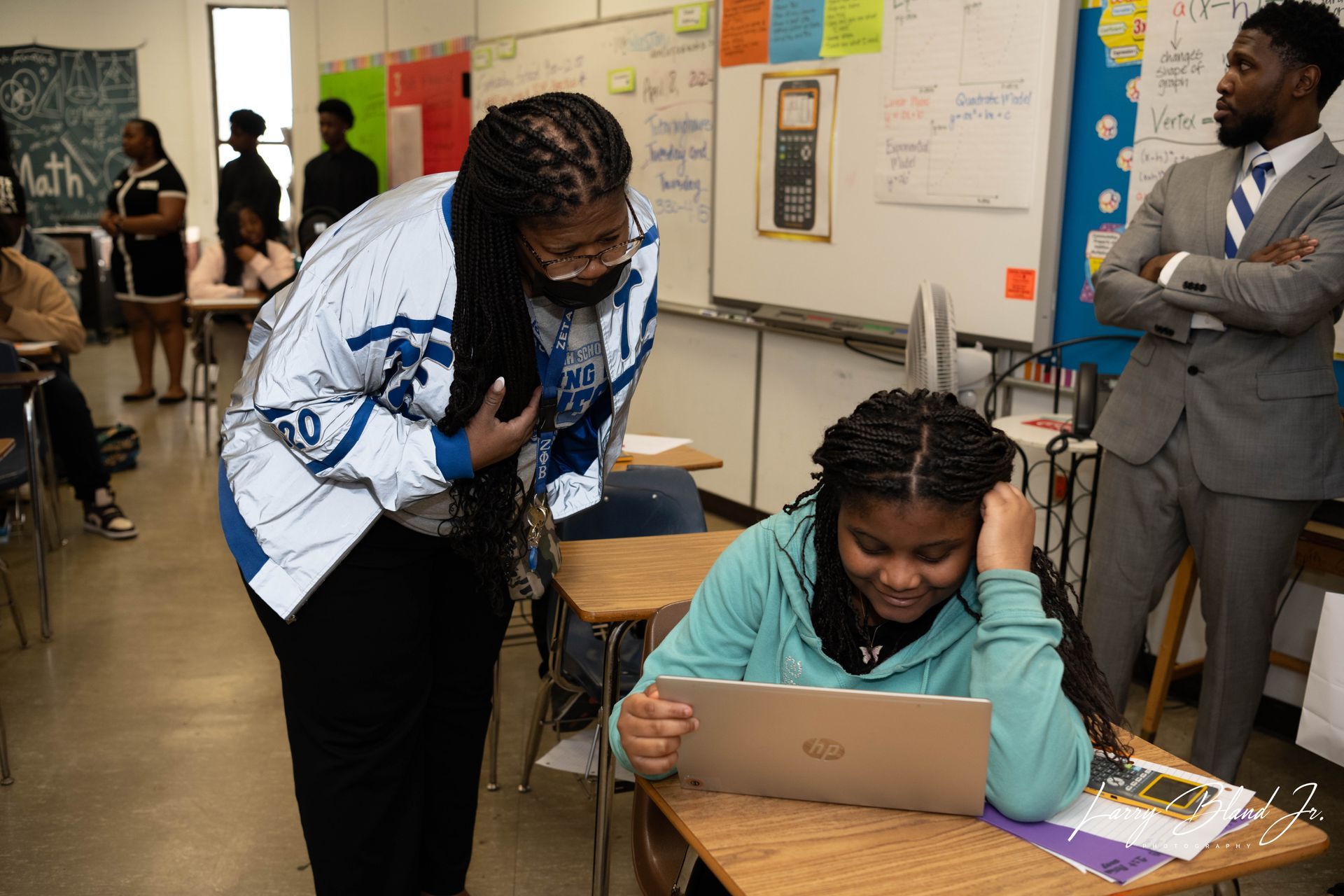 Teacher helping student with laptop in classroom; another man watches.