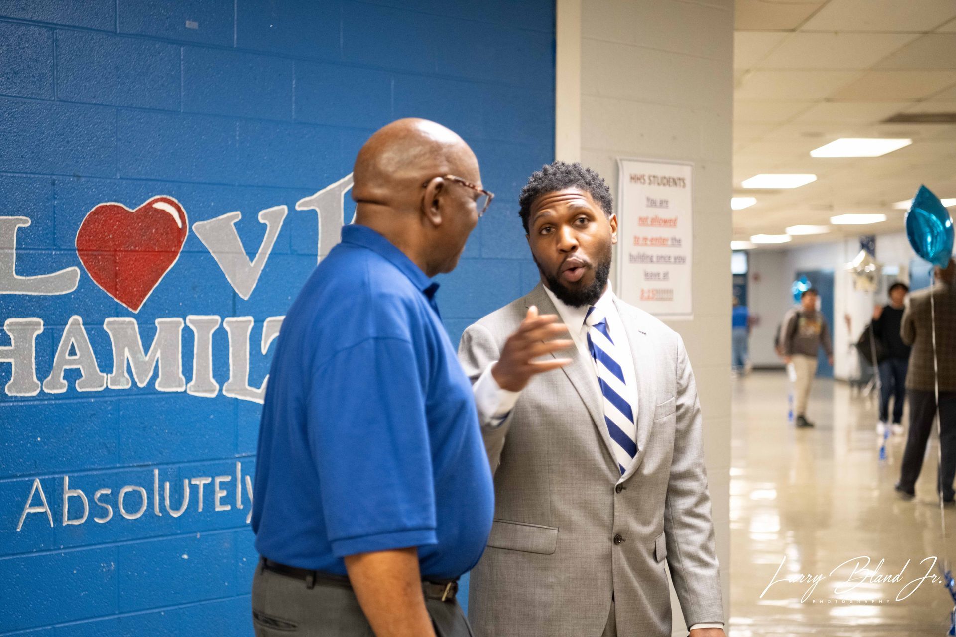 Two men talking in a hallway, one gesturing. Blue wall with 
