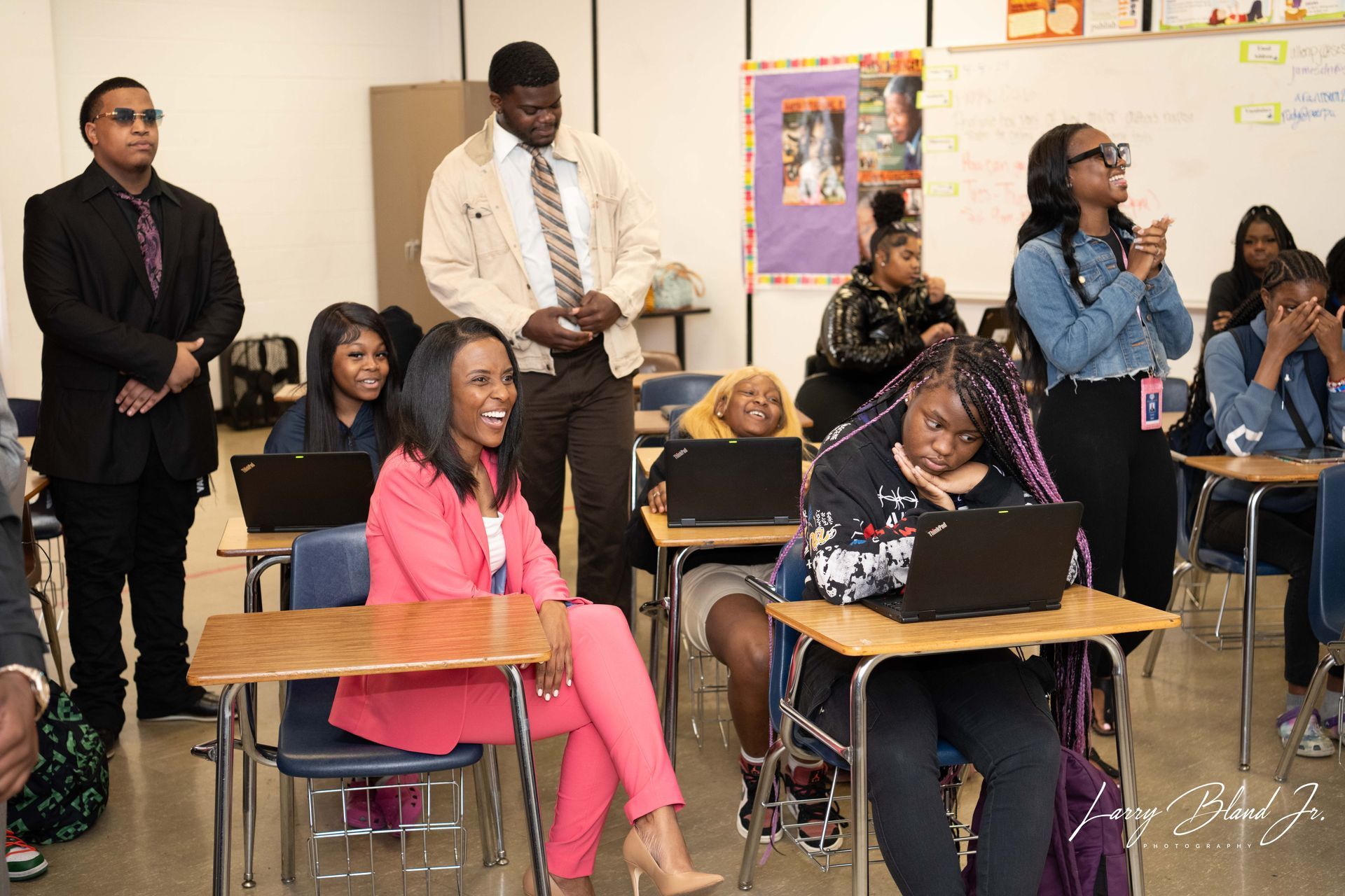 People in a classroom, some using laptops, smiling. A woman in pink, a man with a tie.