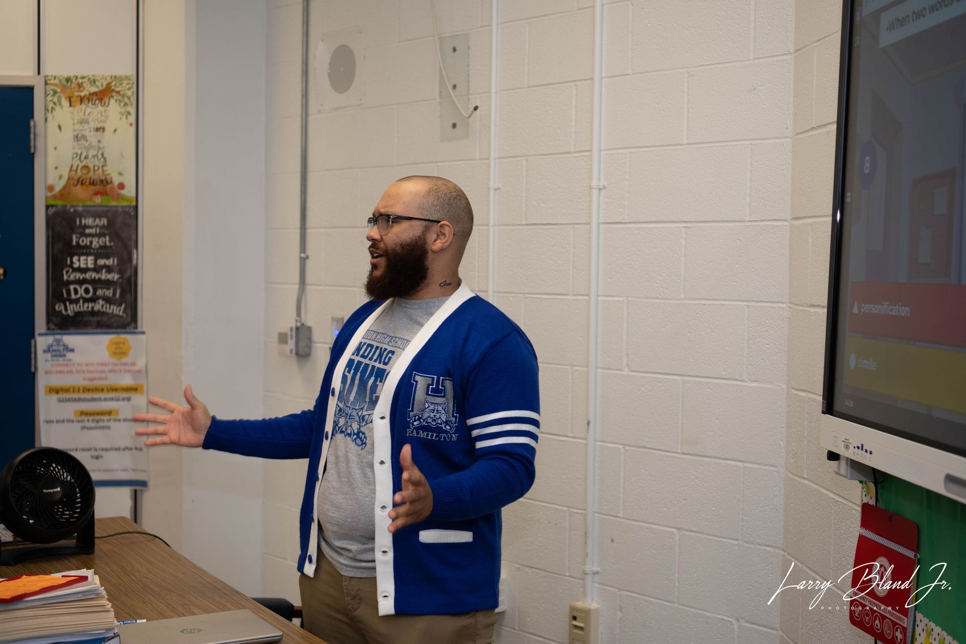 Man with a beard in a blue cardigan gestures during a presentation in a classroom.