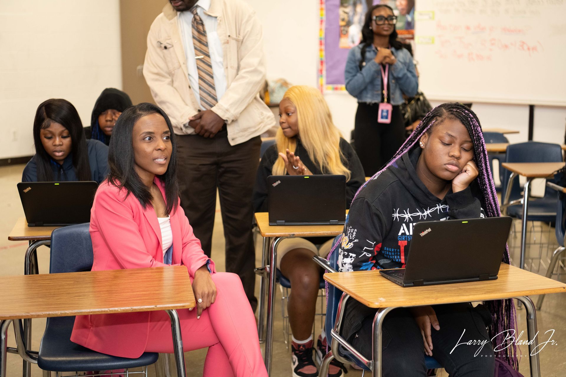 Students and adults in a classroom, some using laptops. A woman in a pink suit looks on.