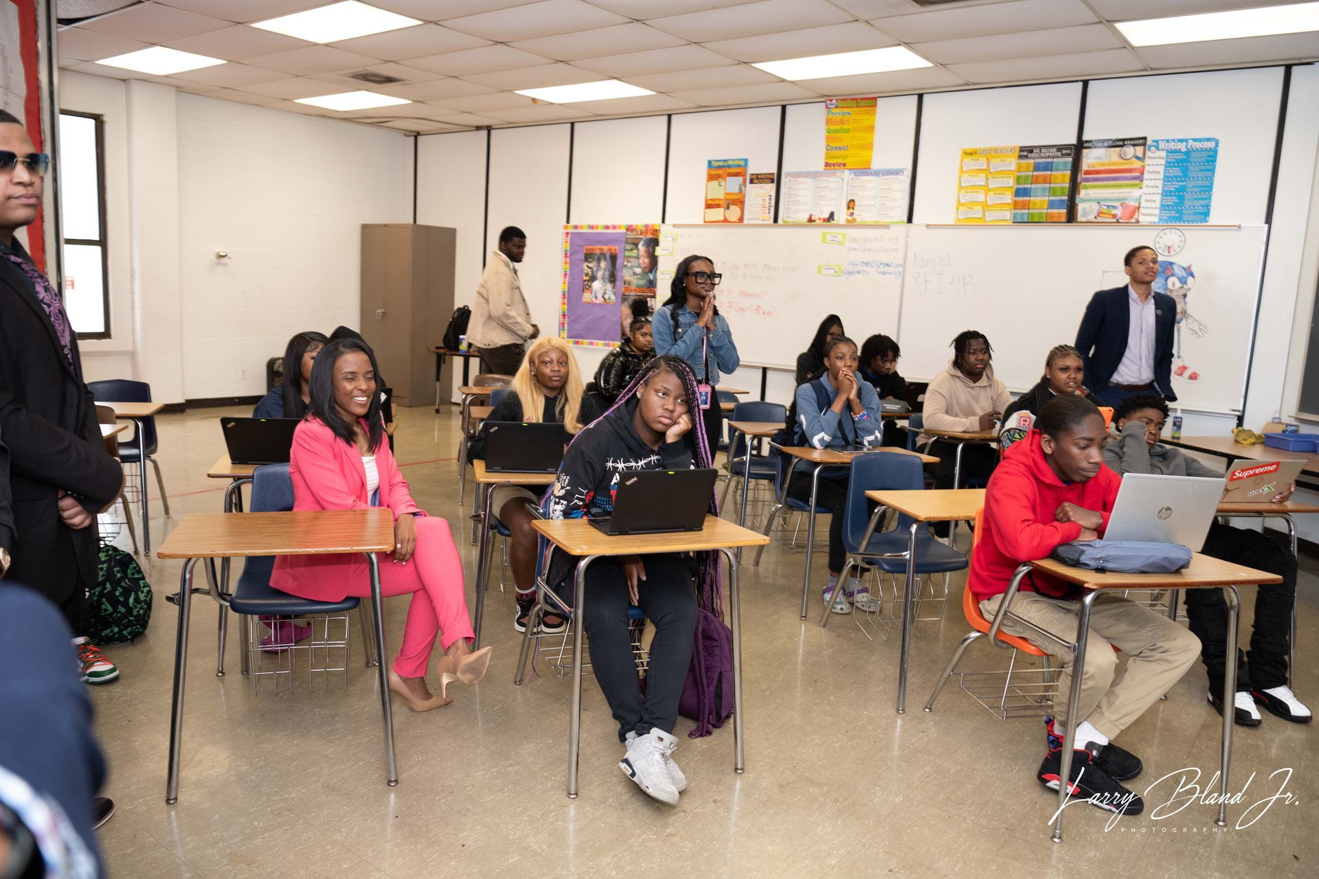 Students in classroom at desks, some using laptops. Adults stand nearby.