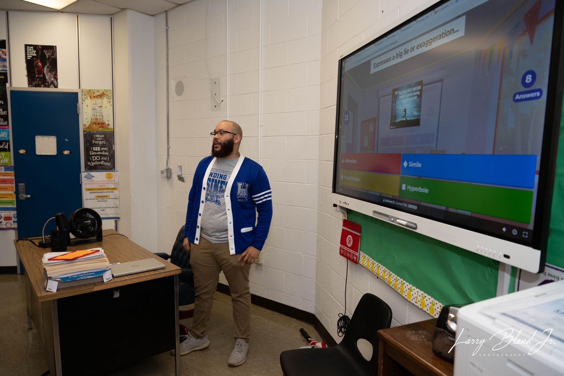 A man stands in a classroom, looking at a screen displaying a quiz. He's wearing a cardigan, and there is a desk nearby.