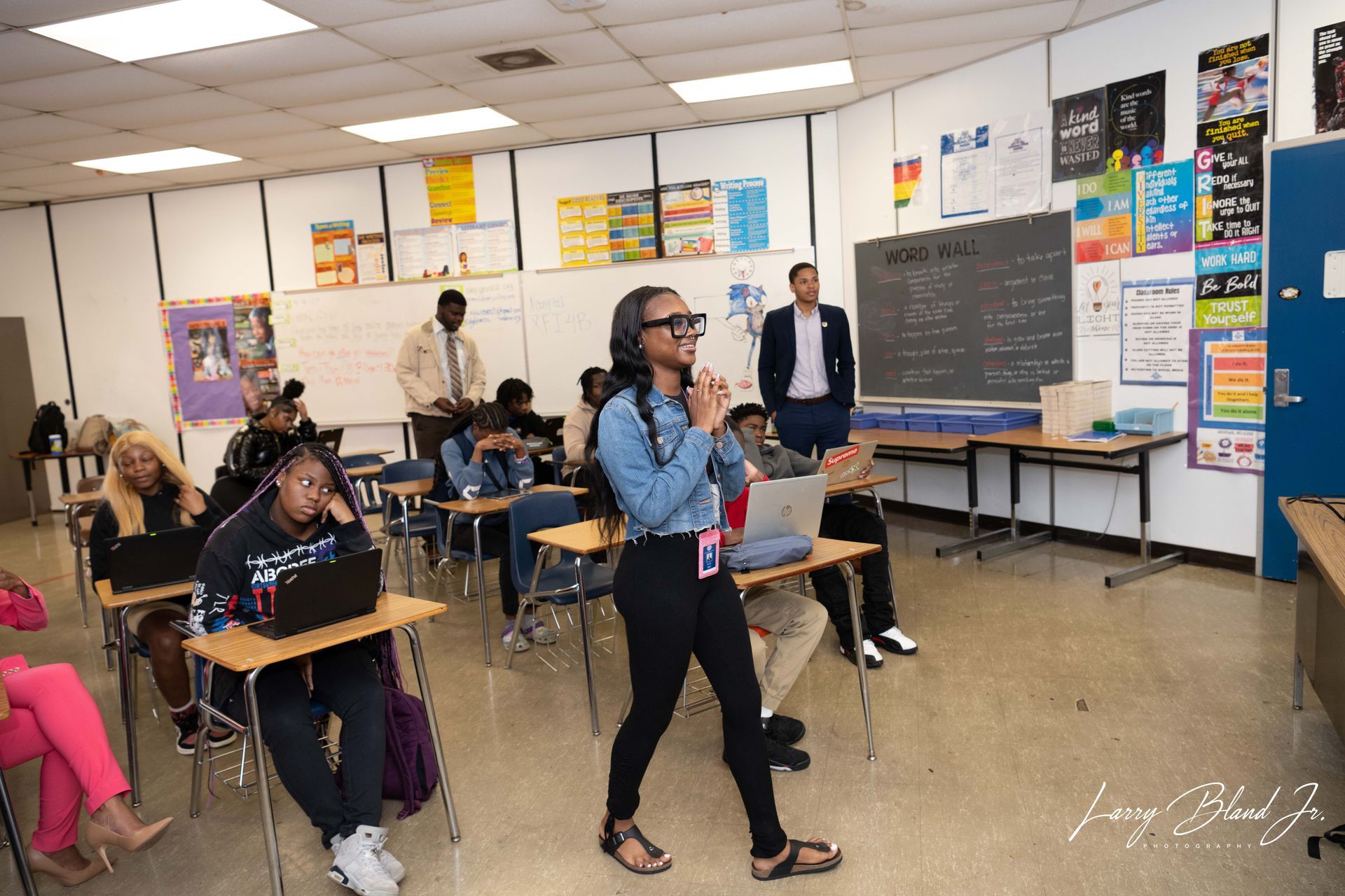 Students in a classroom, one with 3D glasses gesturing, others at desks with laptops. Two adults in the background.