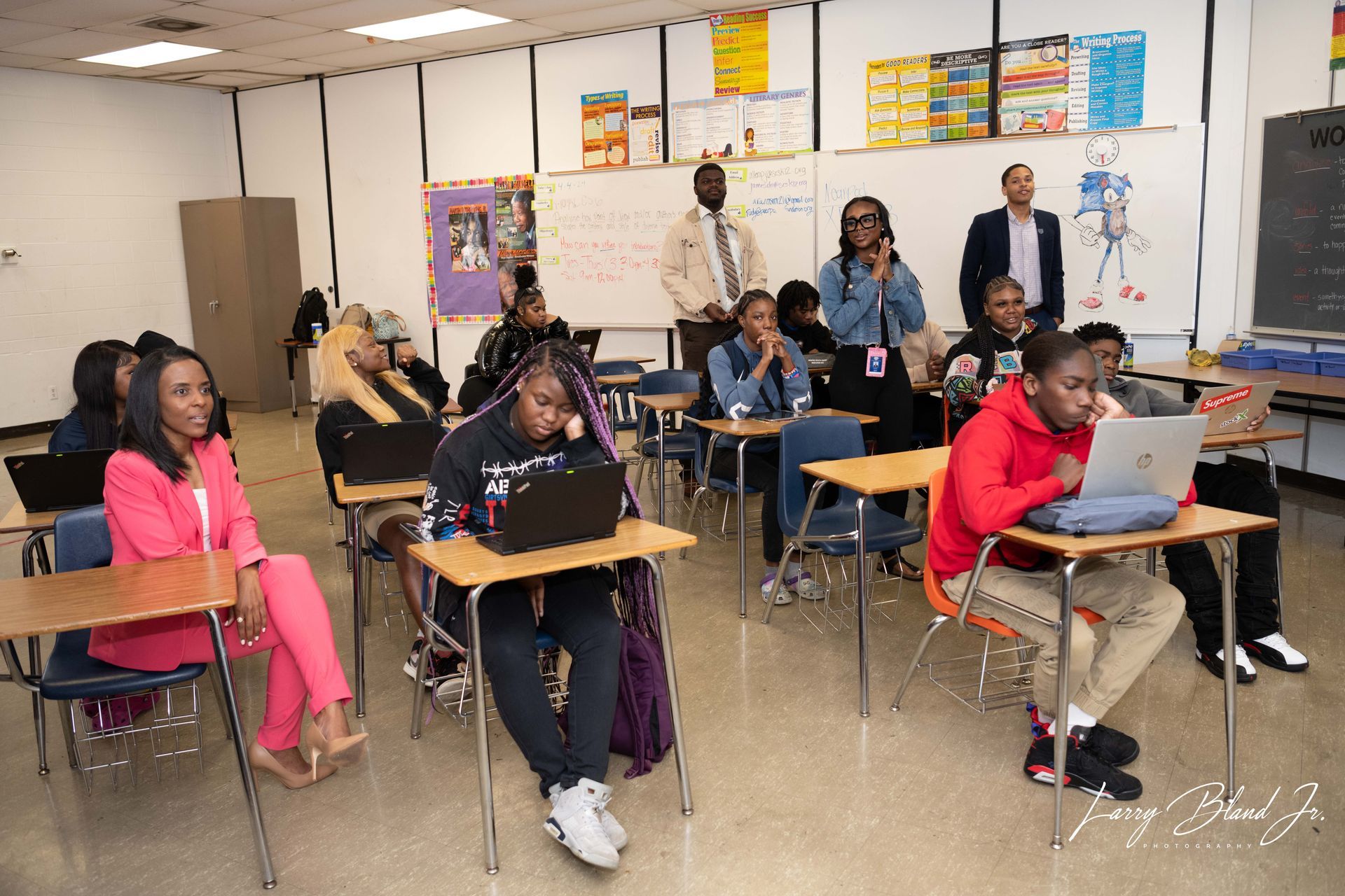 Classroom with students at desks and adults standing. Students use laptops, others look on.