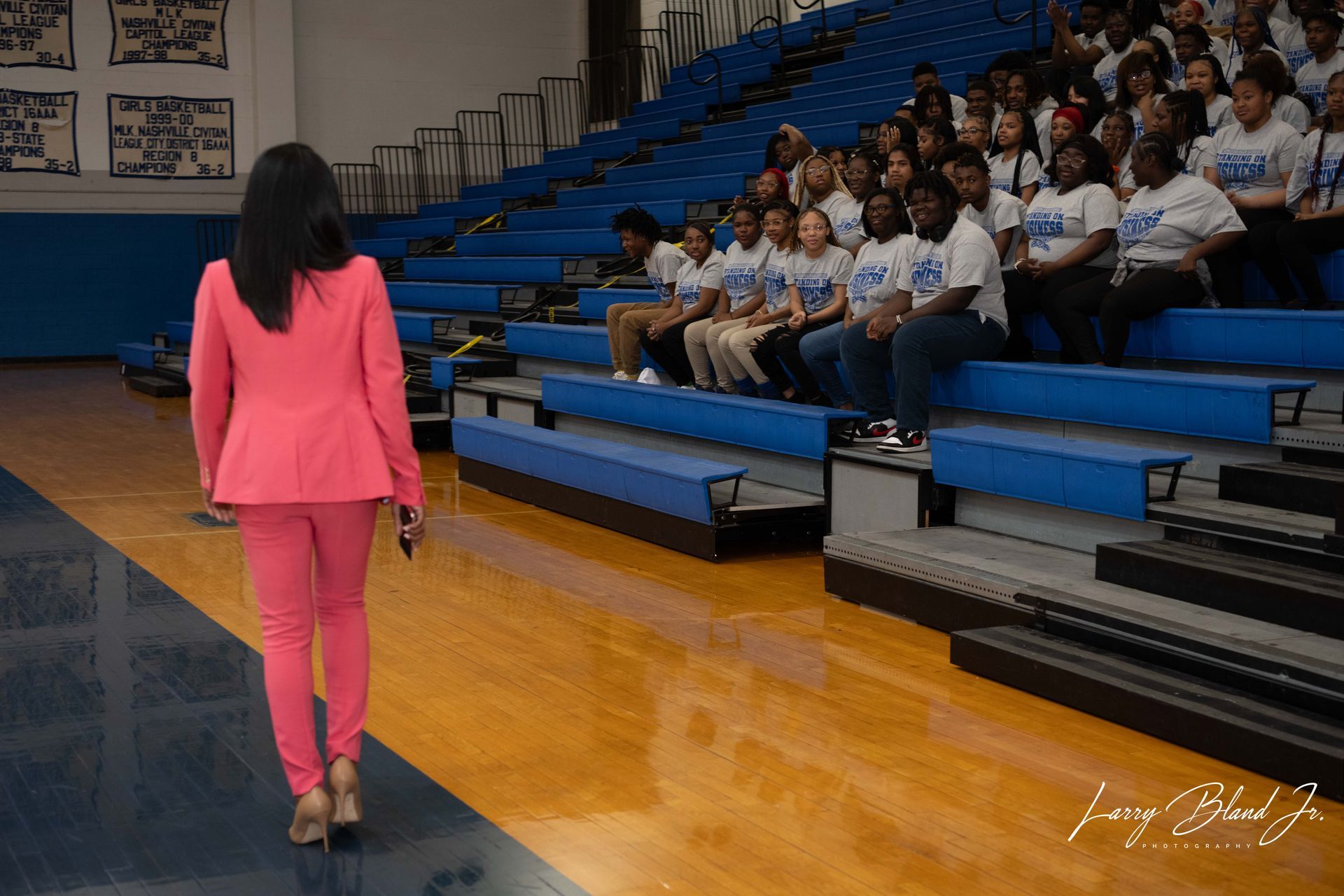 Woman in pink suit walks toward a seated group in a blue-seated gymnasium.
