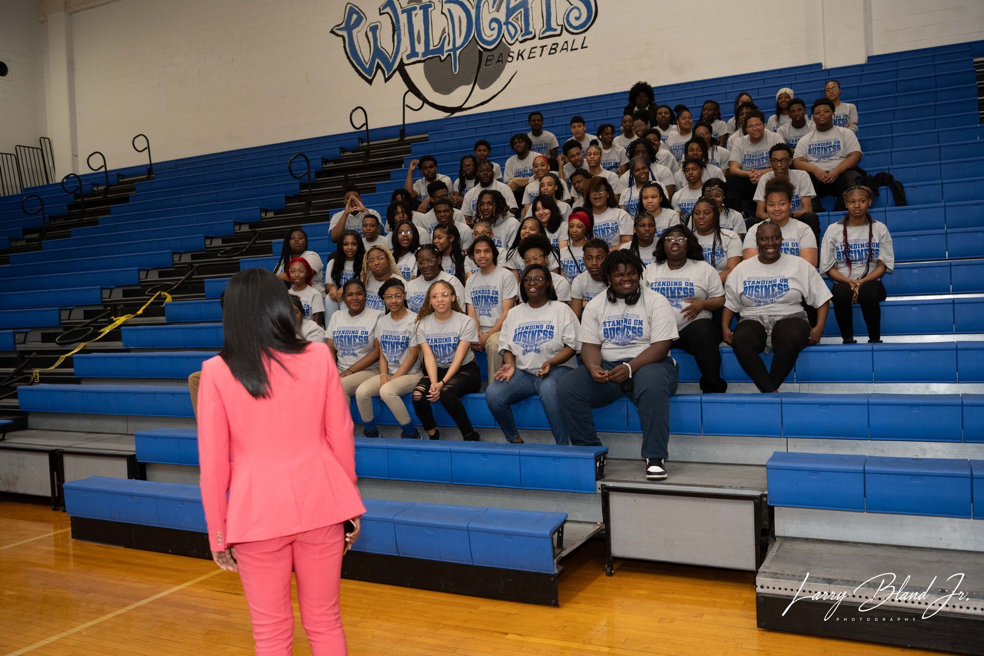 Woman in pink facing group of people in blue shirts on bleachers in gym; 
