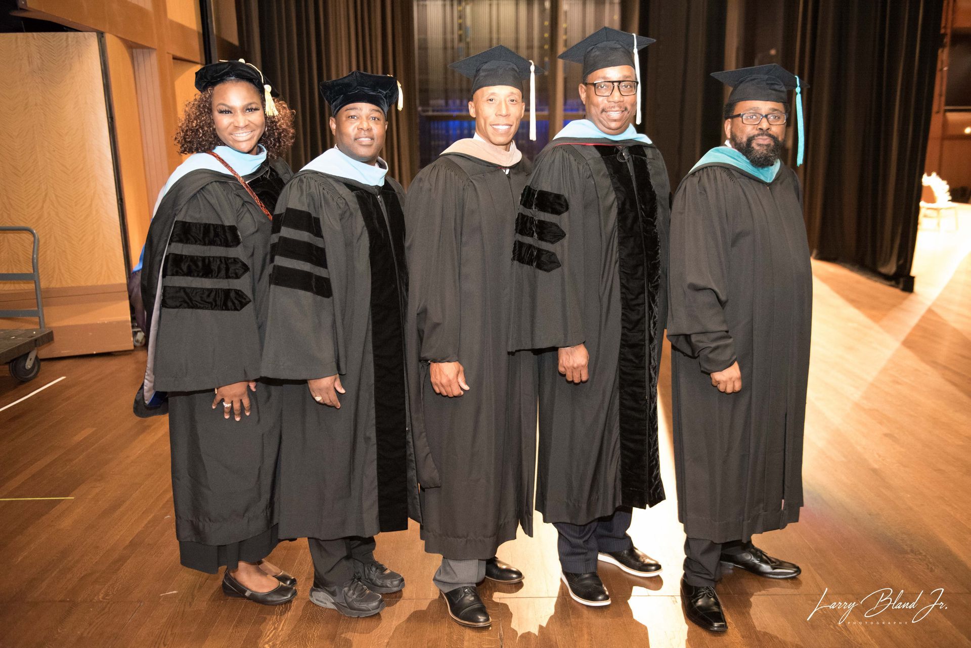 Five people in graduation gowns pose on a wooden floor, smiling.