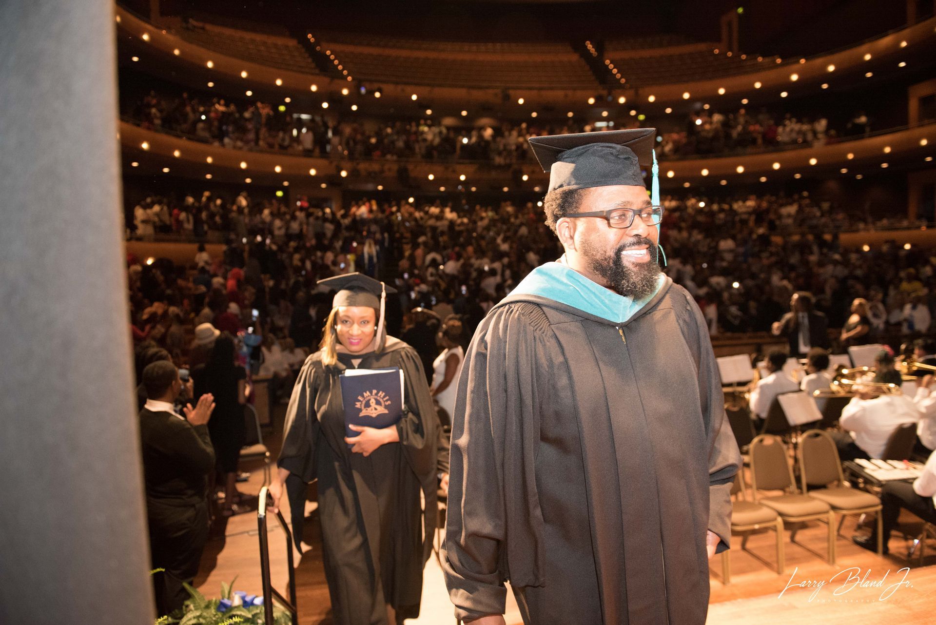Graduates in cap and gowns walk across stage at a ceremony in a packed auditorium, smiling.