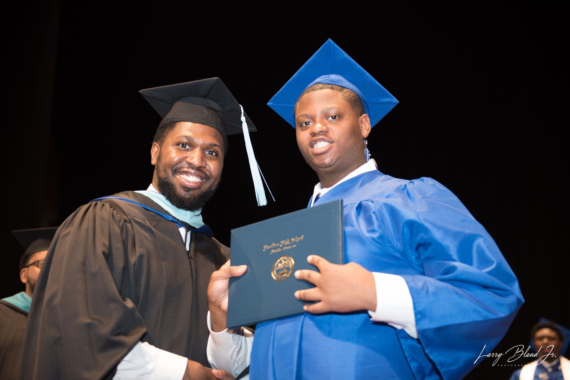 Man in blue graduation gown receives diploma from man in black robe.