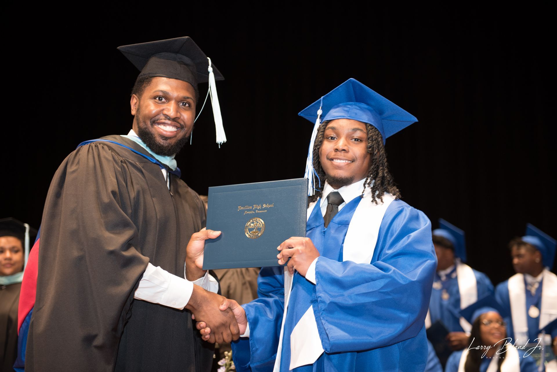 Graduate receiving diploma from a robed person on stage, blue and black gowns, handshake.