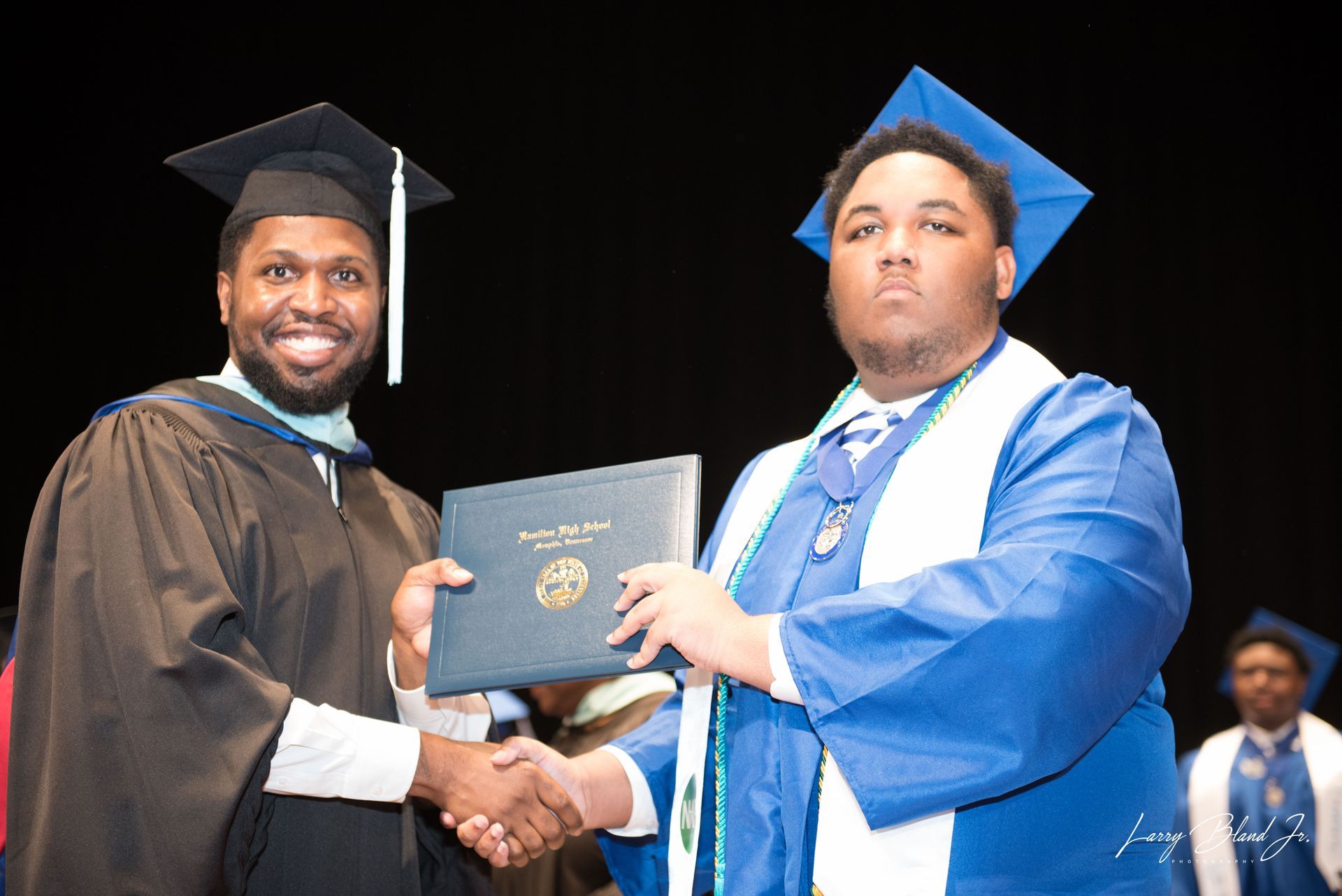 Person in graduation gown presents diploma to another at a ceremony, shaking hands.