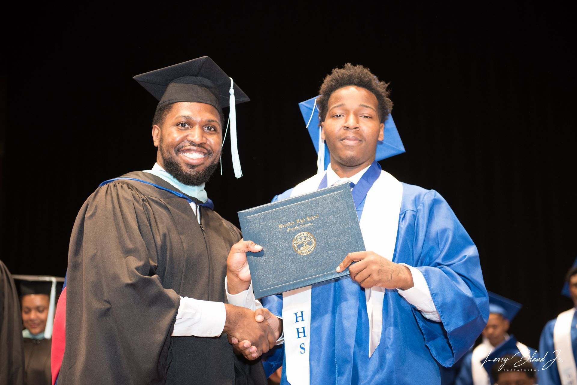 A person in a graduation gown hands a diploma to a student in a blue gown; both are smiling.