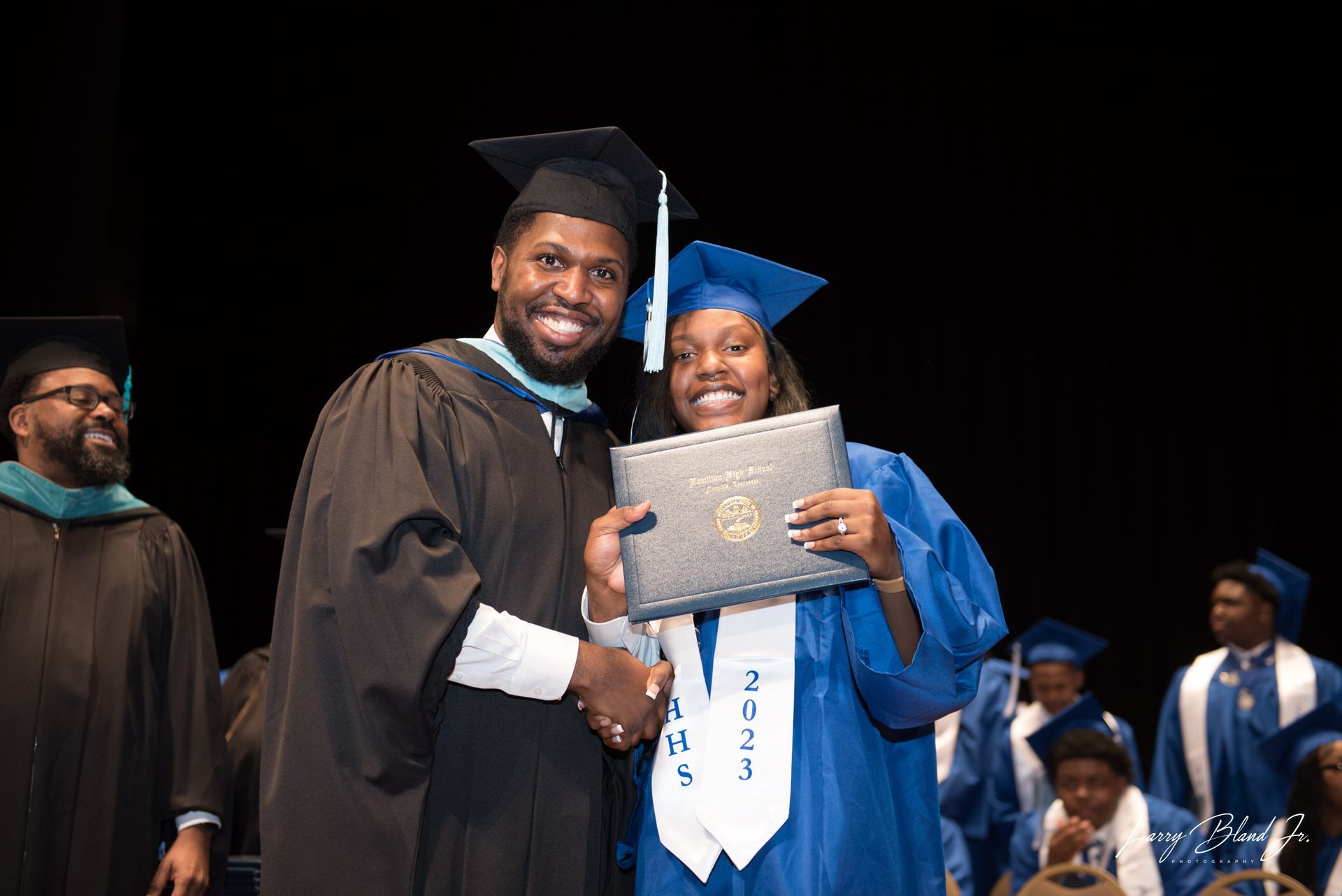 Graduation ceremony: Man hands diploma to smiling person in blue gown and stole, on stage.
