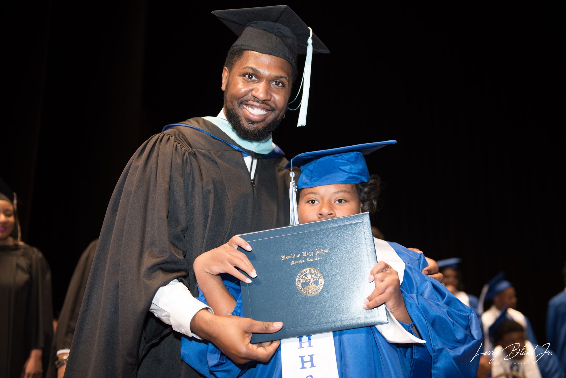 Man in graduation garb presents diploma to student in a blue gown; both smile.