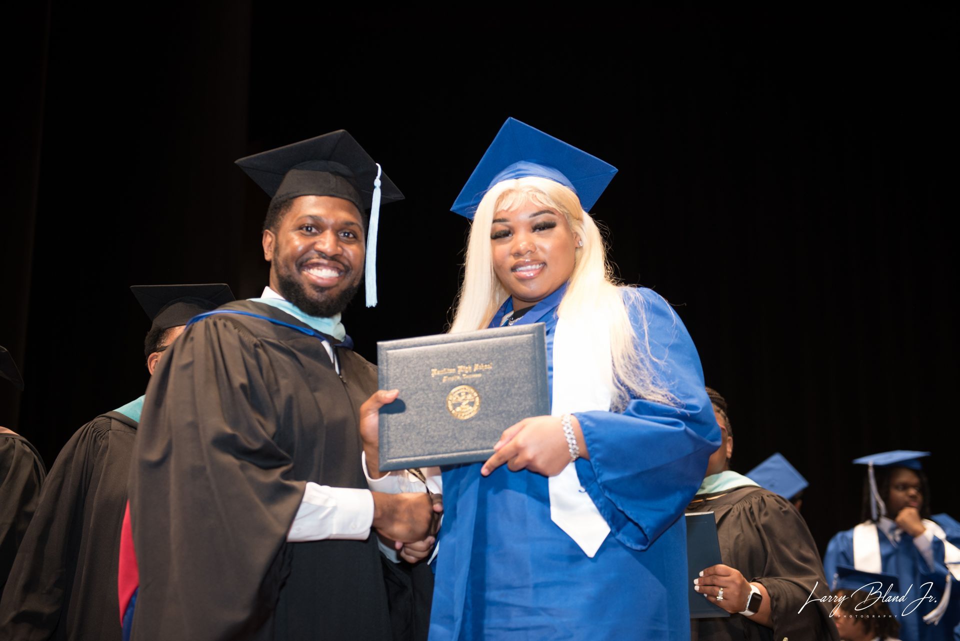 Graduation ceremony: Man in black robe hands diploma to person in blue gown and cap.