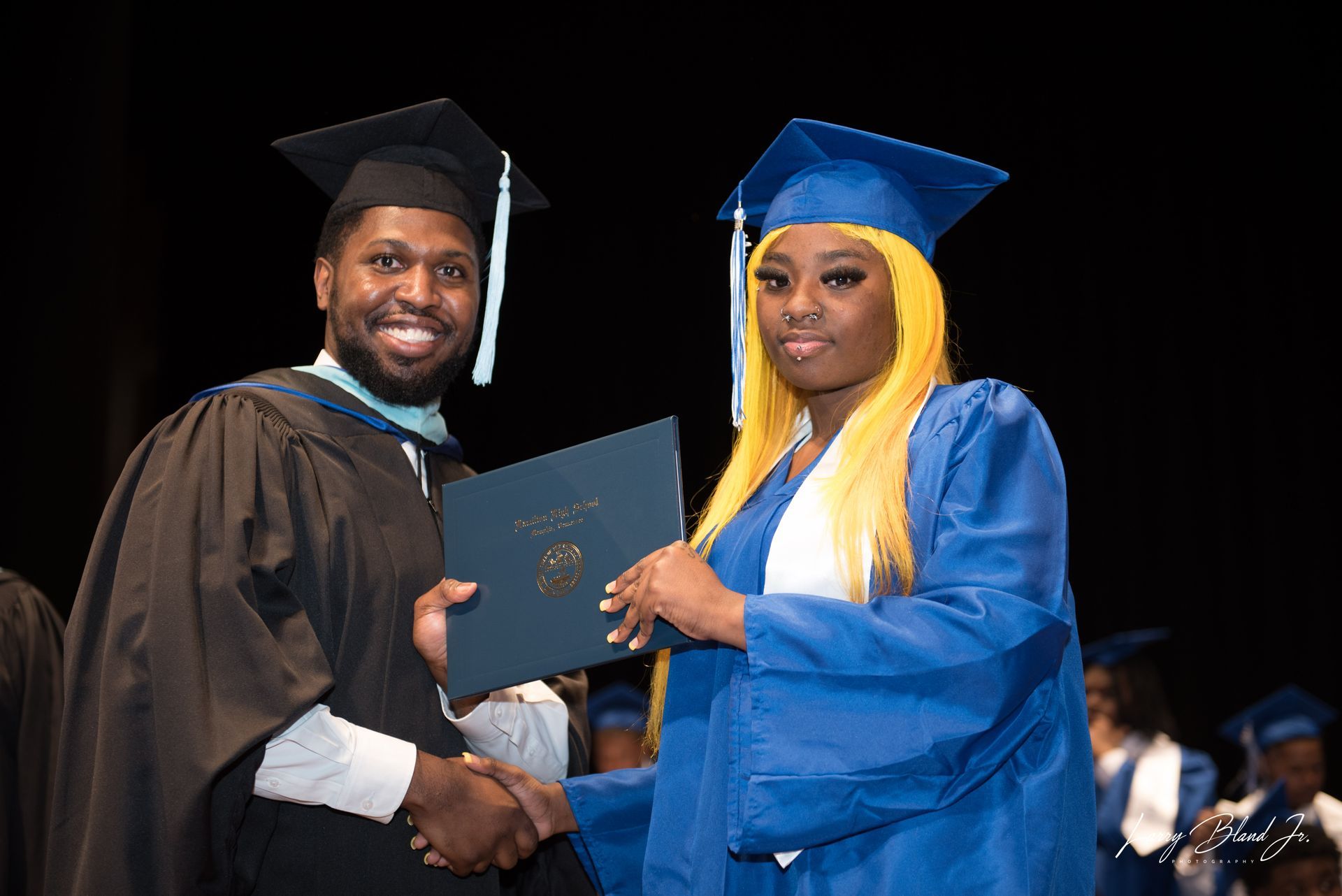 Person in black robe handing diploma to person in blue gown; graduation ceremony.