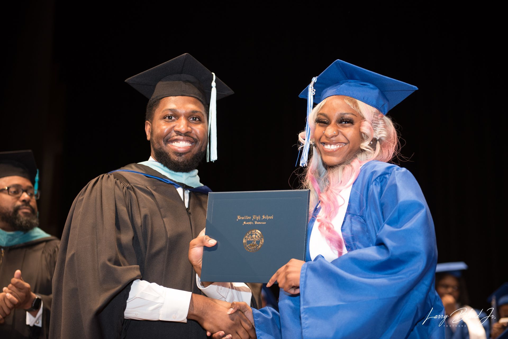 Man in academic regalia hands diploma to smiling person in blue cap and gown.