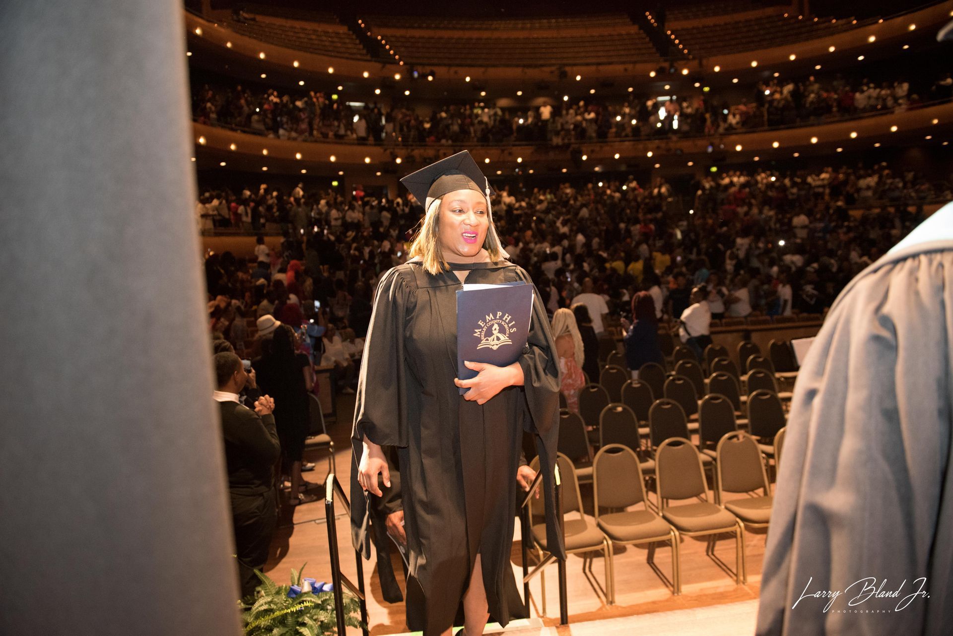 Person in graduation attire walks across a stage, holding a diploma, with a crowd in the background.