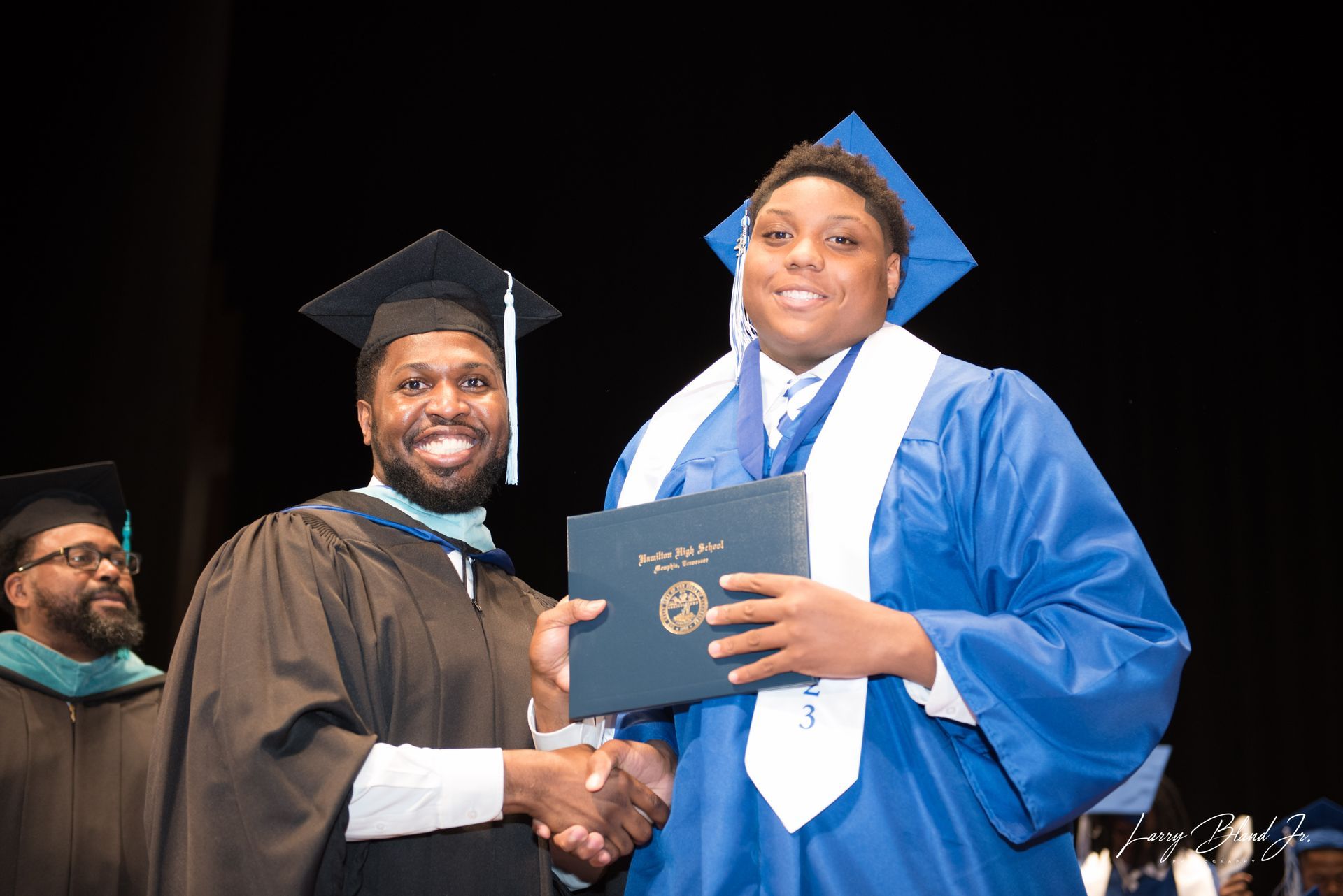 Man in black robe shakes hands with graduate in blue gown, holding diploma.