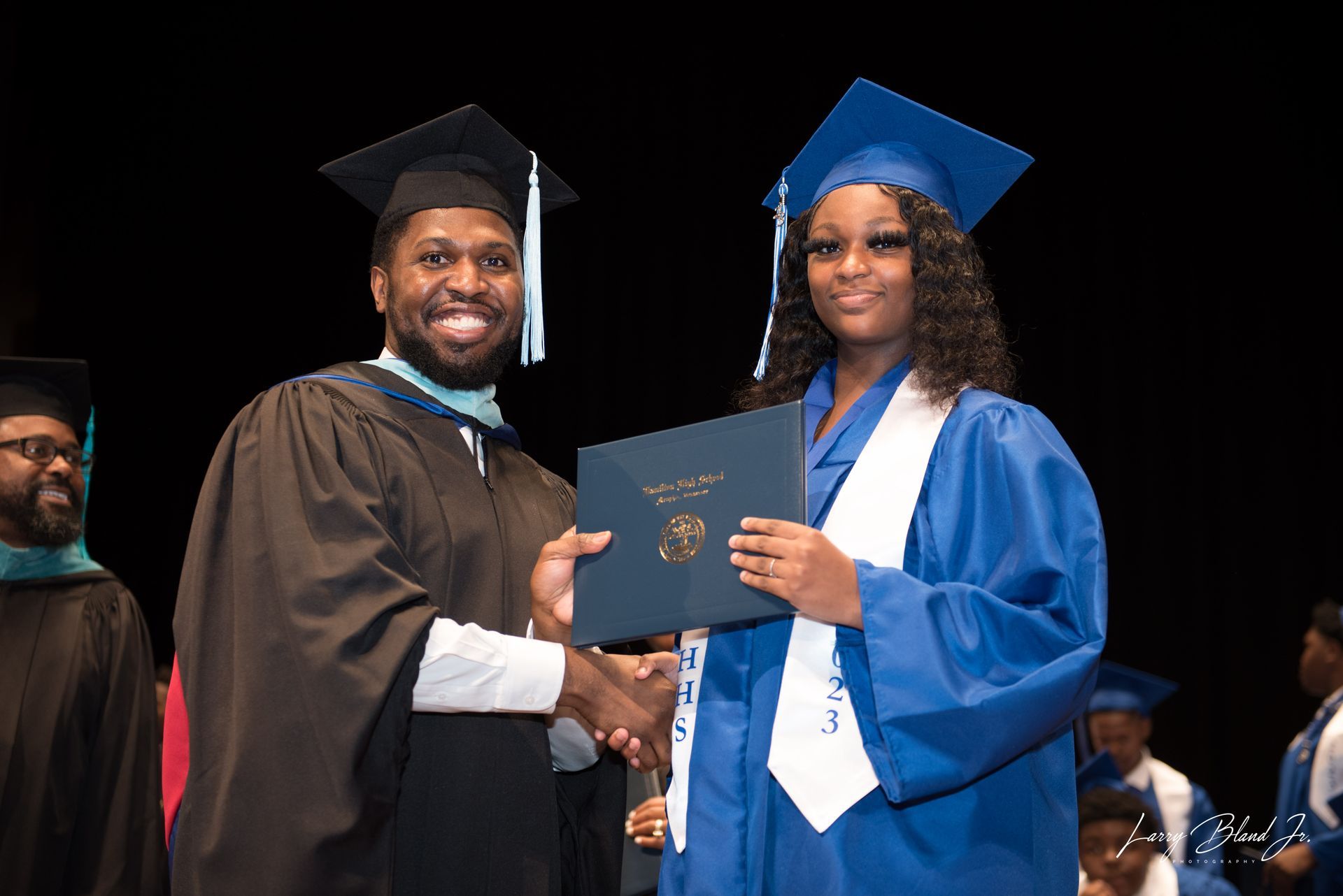 Graduate in blue gown receives diploma from robed person, stage background.