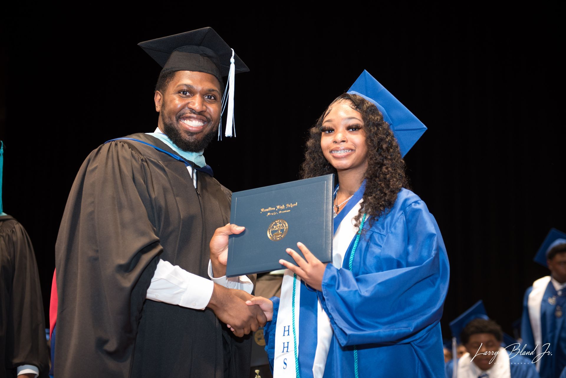 Man in academic robe hands diploma to a woman in blue graduation gown on stage.