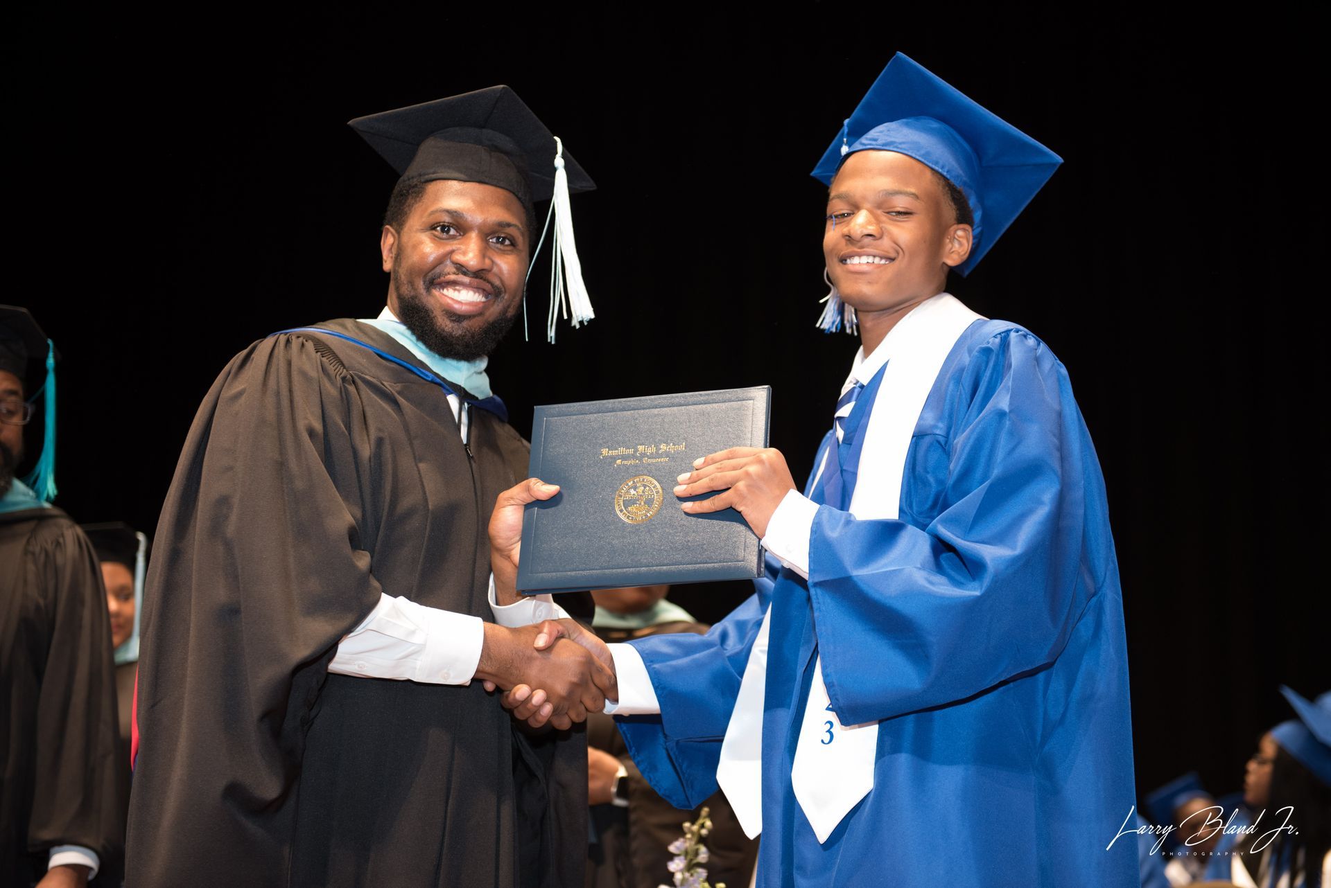 Person in black graduation gown hands diploma to person in blue gown, shaking hands on a stage.