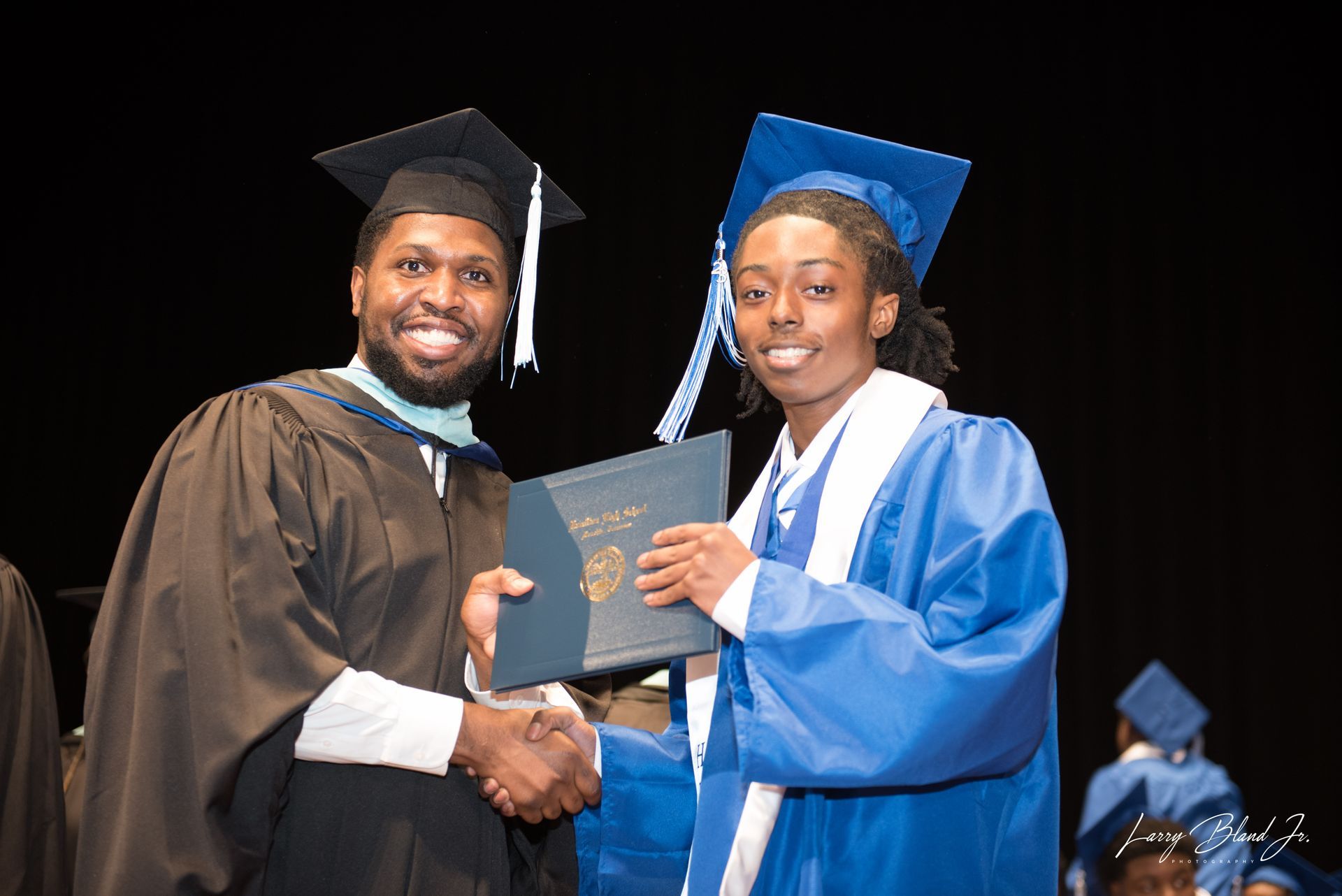 Man in academic regalia shakes hands with a person in blue cap and gown holding a diploma.