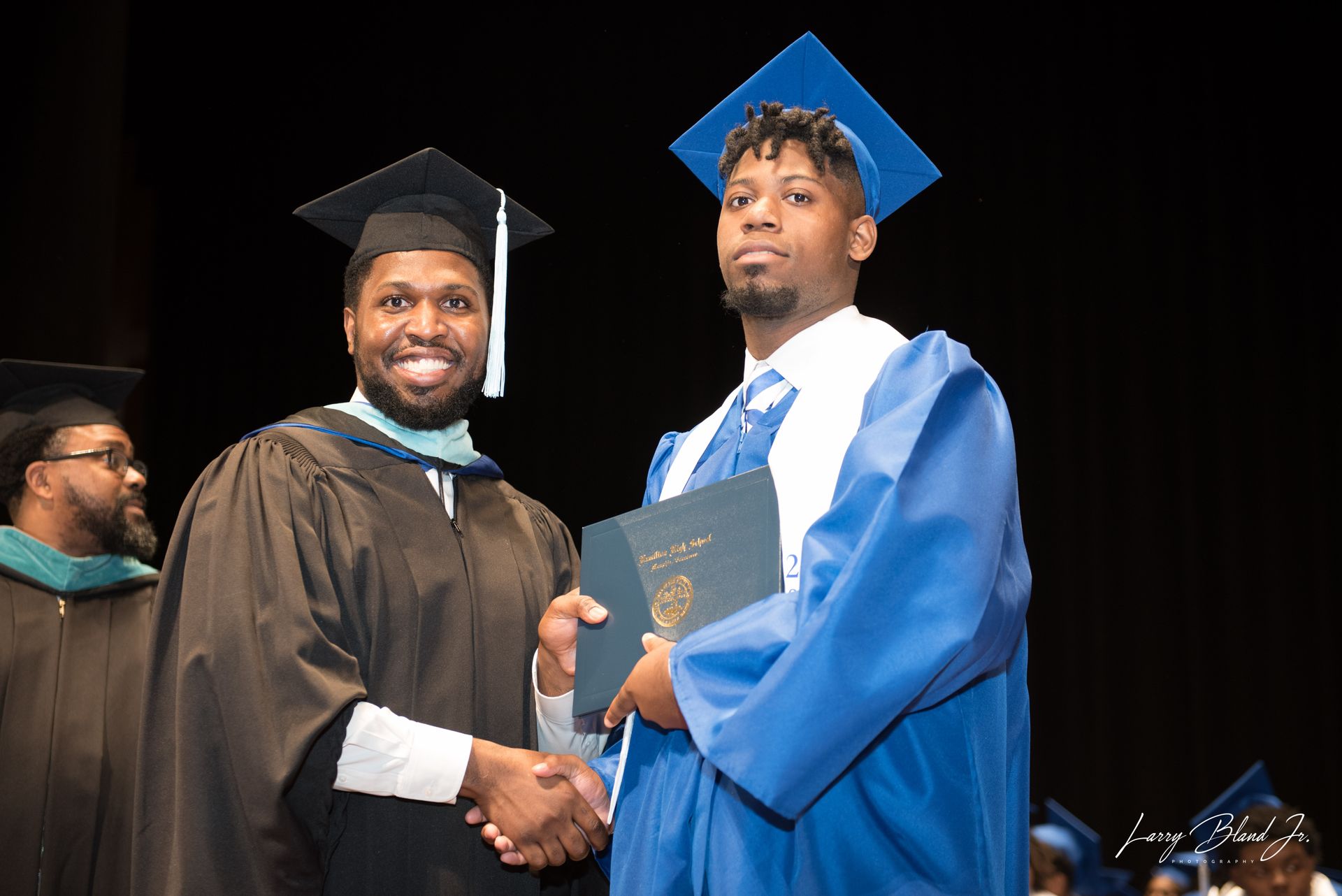 Man in cap and gown shakes hands with graduate in blue gown, diploma in hand. Stage setting.
