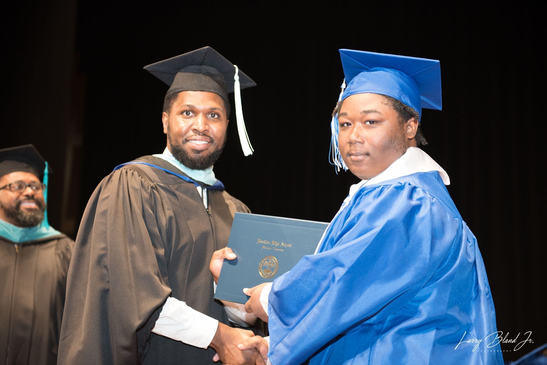 Man in black cap and gown hands a diploma to a person in a blue cap and gown on a stage.