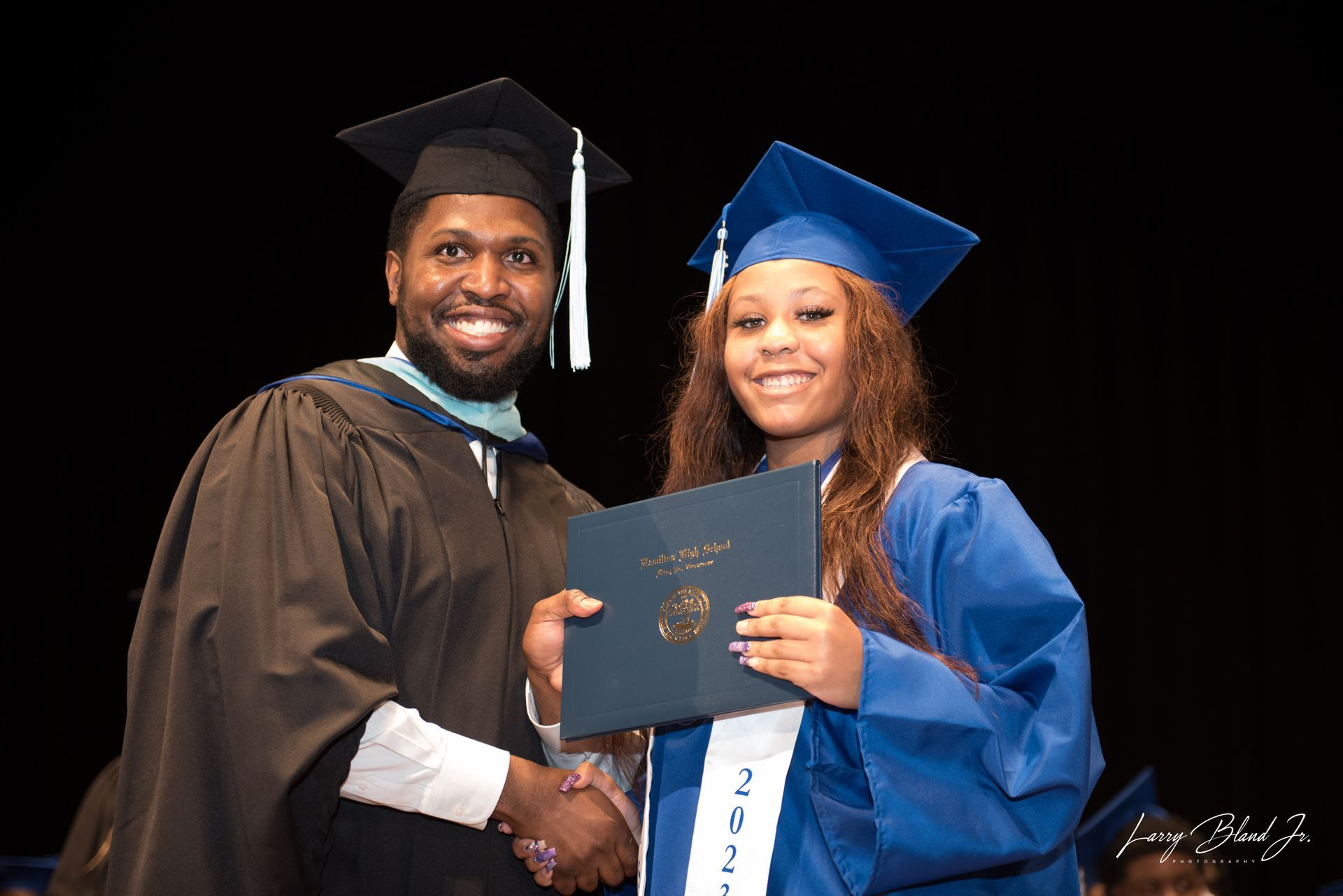 A person in a blue graduation gown receives a diploma from a person in a black gown, both smiling, indoors.