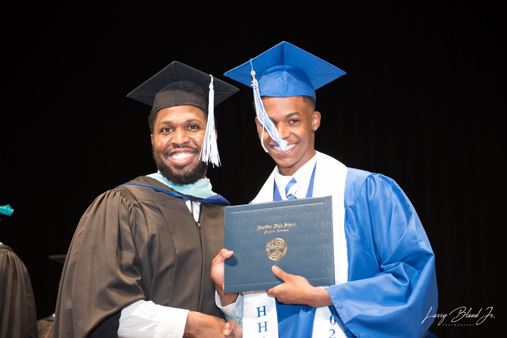 A graduate in blue robe receives a diploma from a robed person on stage.