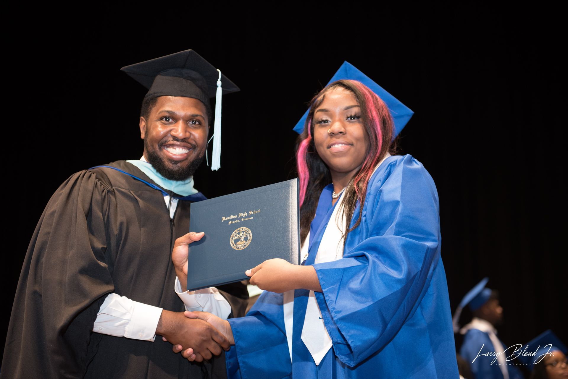 Man in academic robe hands diploma to graduate in blue gown.