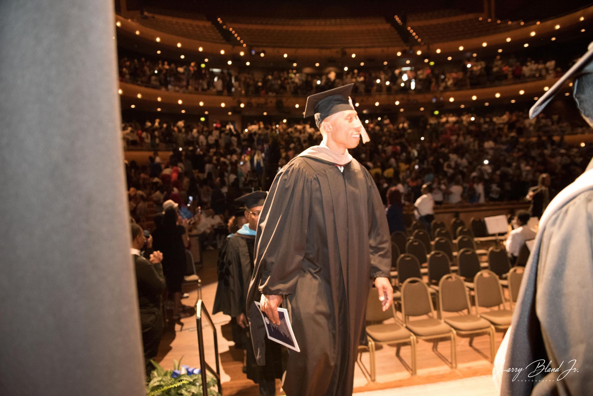 Graduation ceremony with graduates in black gowns, walking toward seats, and an audience in the background.