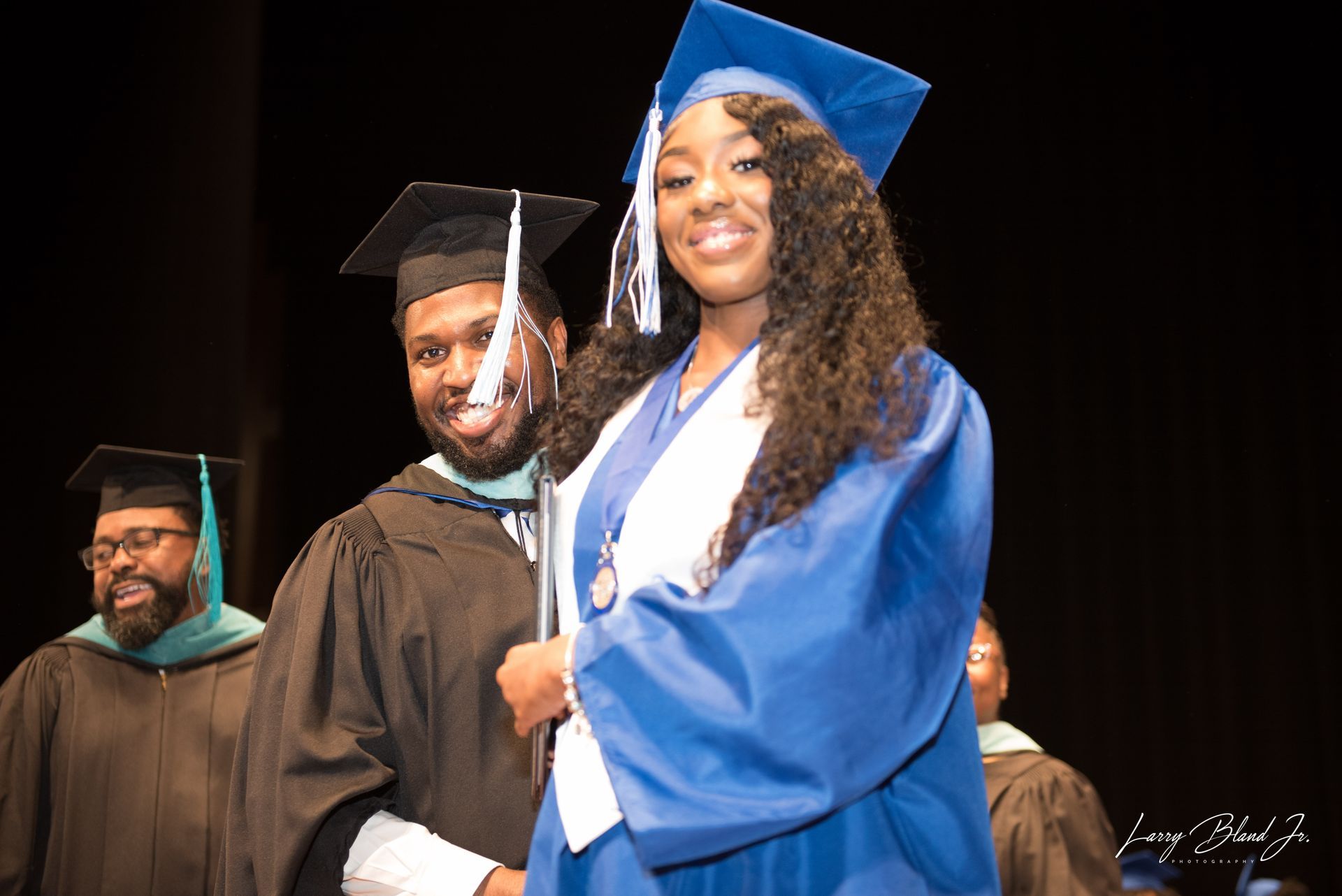 Graduates in blue and black gowns smile at a ceremony, one holds a diploma, indoors.