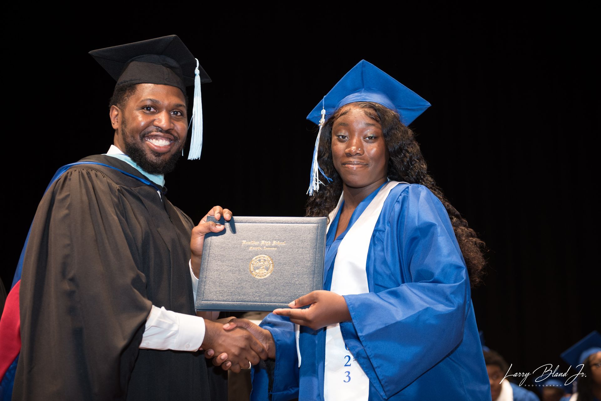Man in black graduation attire shakes hands with woman in blue gown, presenting a diploma on stage.