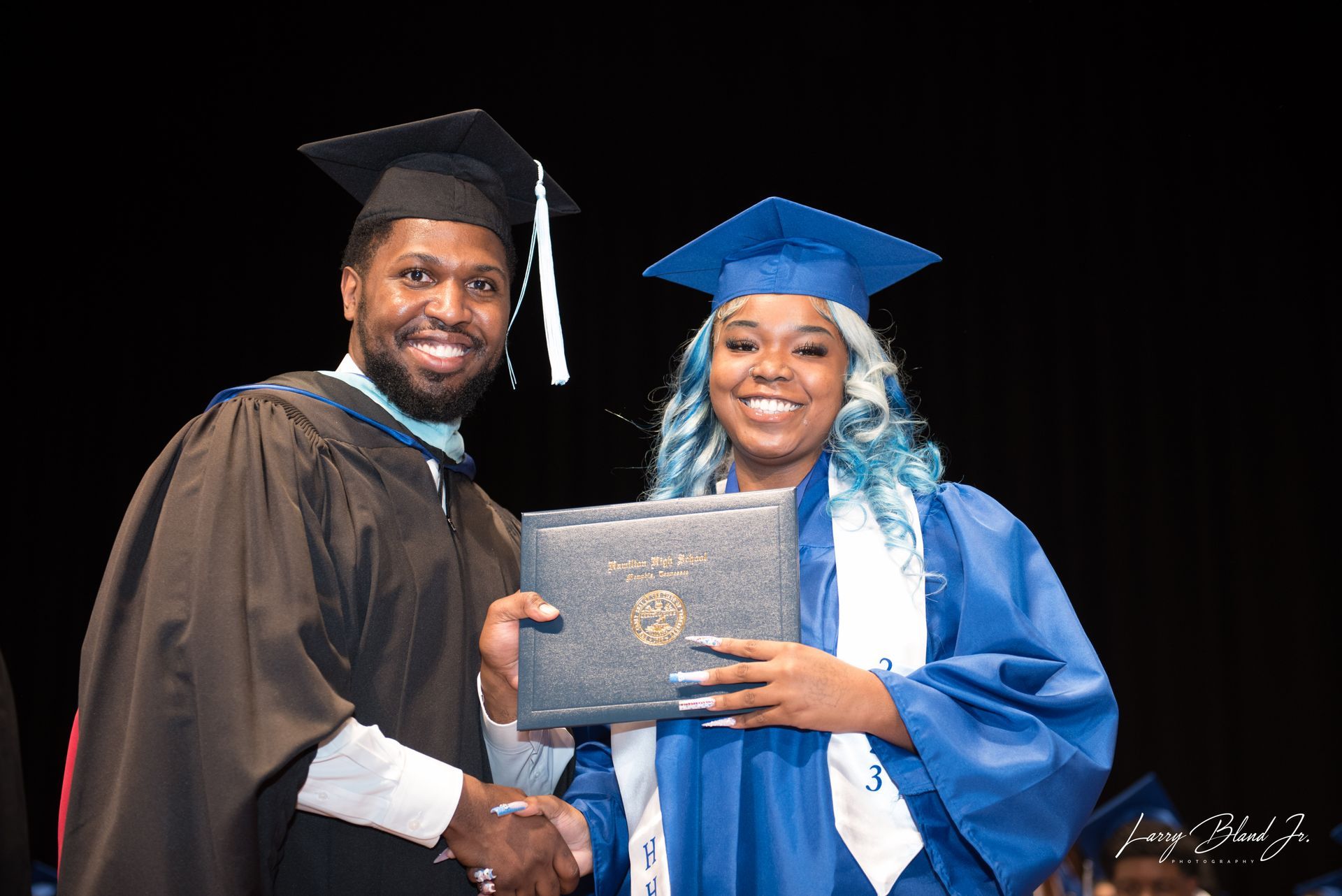 Person in graduation gown hands diploma to another smiling graduate.