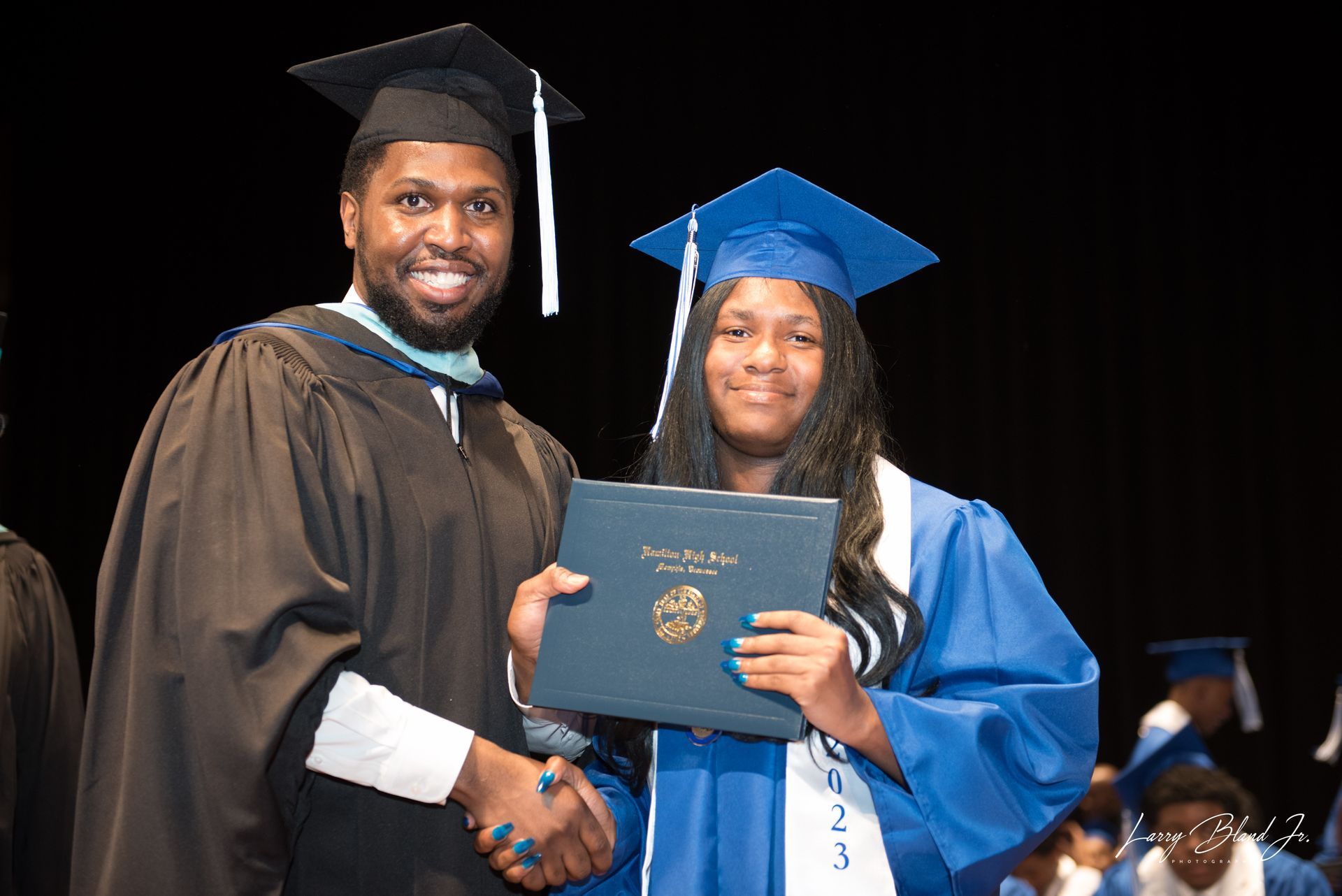 Person in graduation gown shaking hands with a graduate in blue gown, holding a diploma.