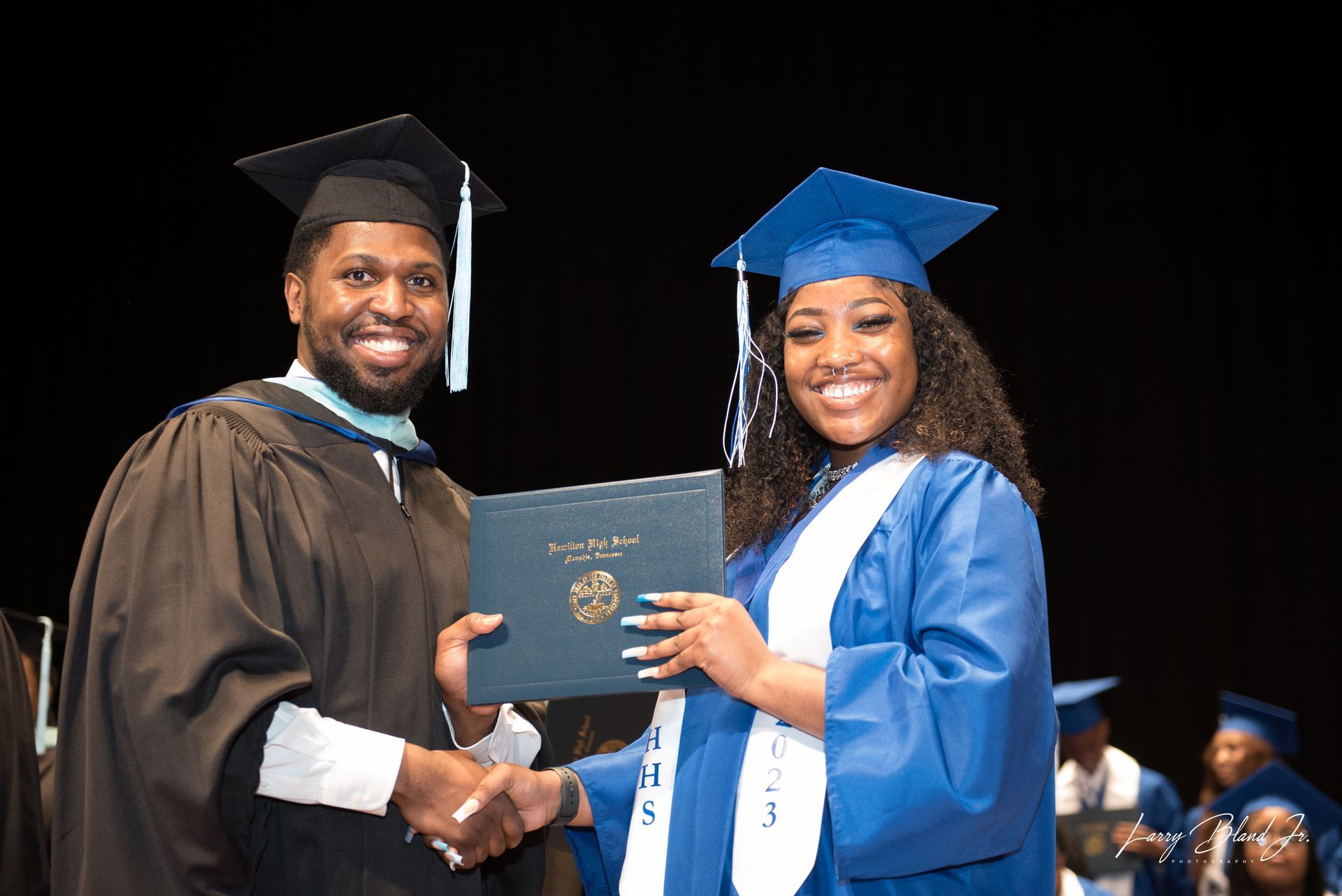 Man in black robe hands diploma to graduate in blue gown.