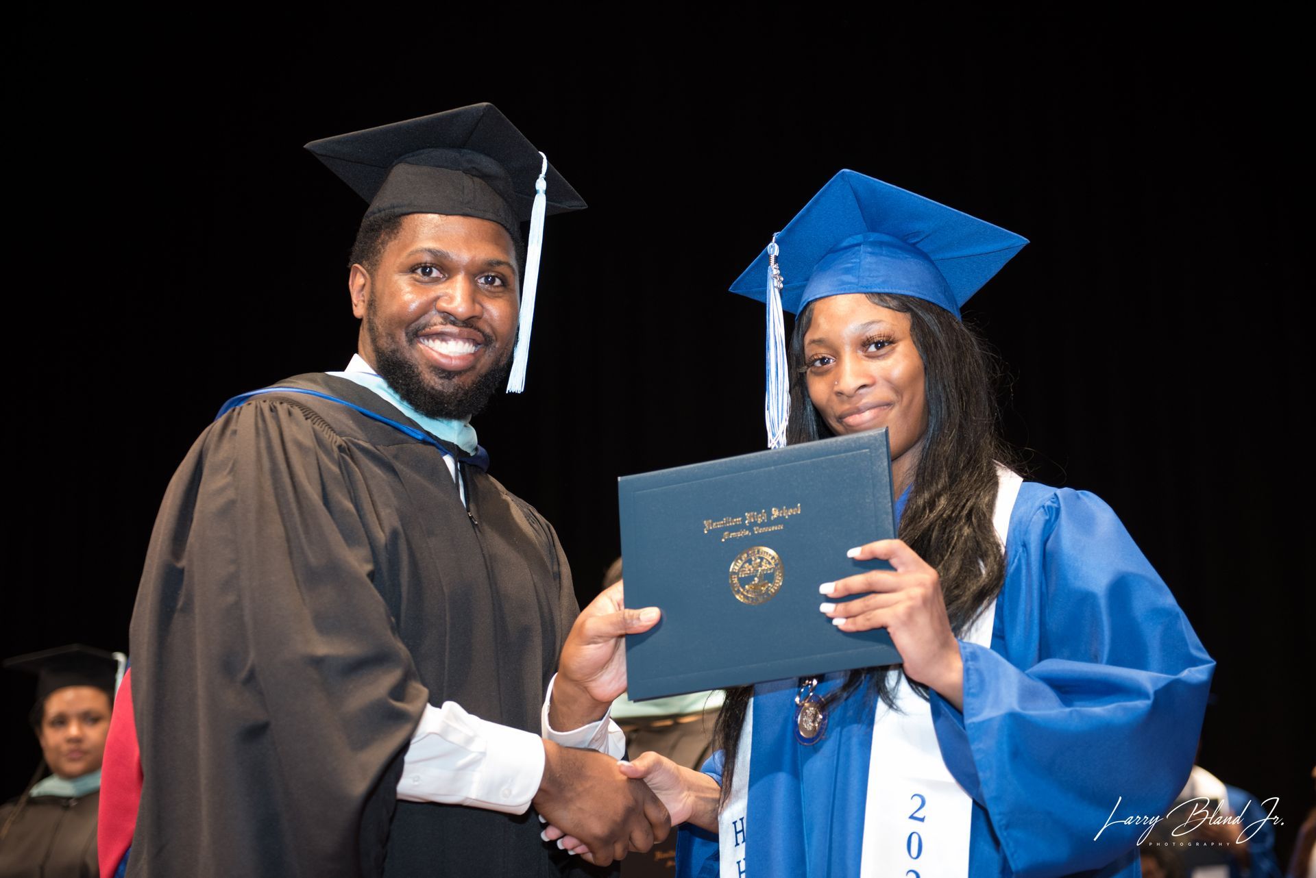 Man in graduation gown hands diploma to woman in blue gown.