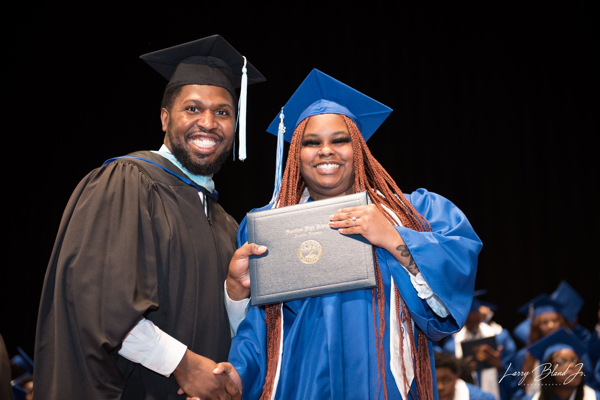 Man in academic robe hands diploma to graduate in blue gown.