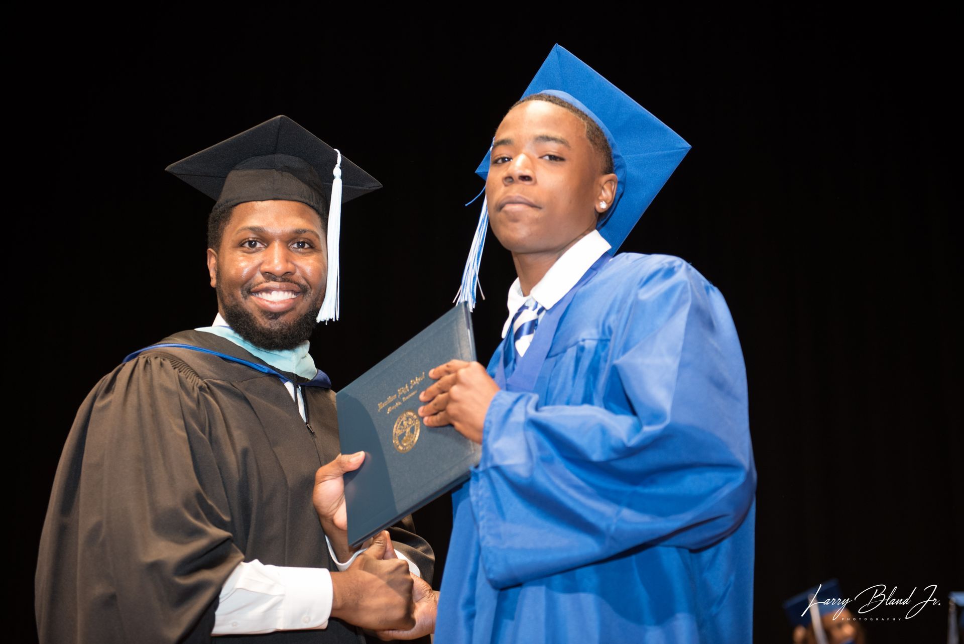 Graduate in blue gown receives diploma from a man in academic regalia, both smiling.
