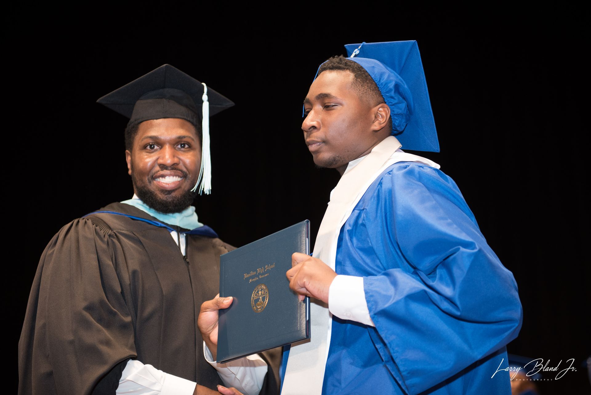 Man in brown graduation gown presents a diploma to a person in a blue gown; both are smiling.