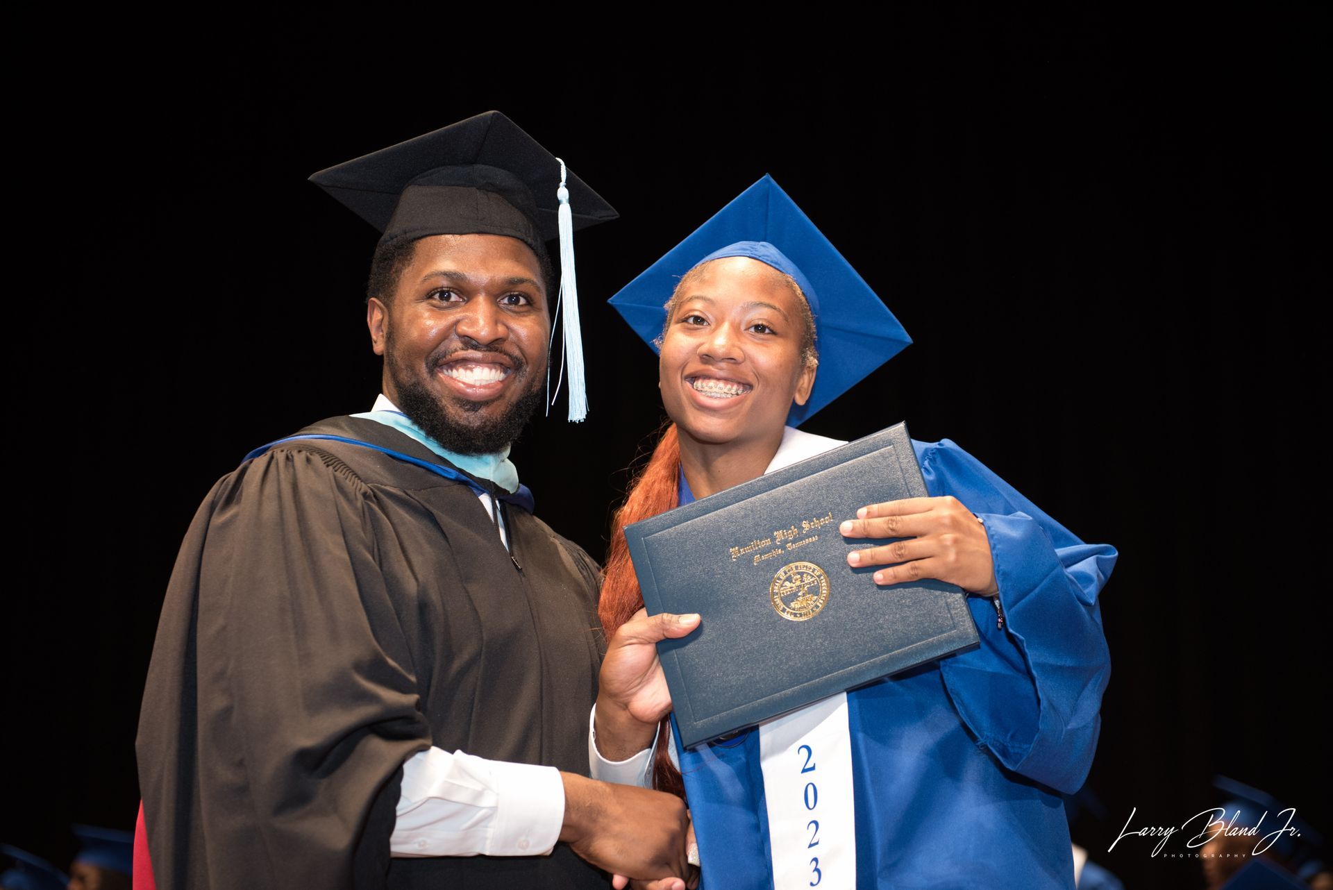 Graduation photo: Person in academic gown shakes hands with graduate in blue cap and gown, holding diploma.