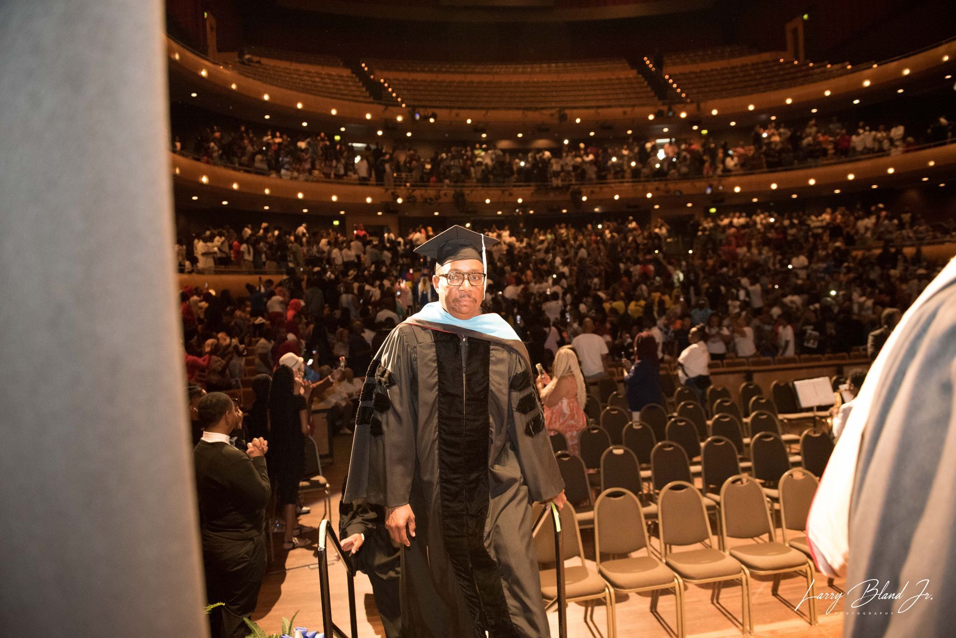 Graduation ceremony: person in gown, walking onstage in theater, audience in background.